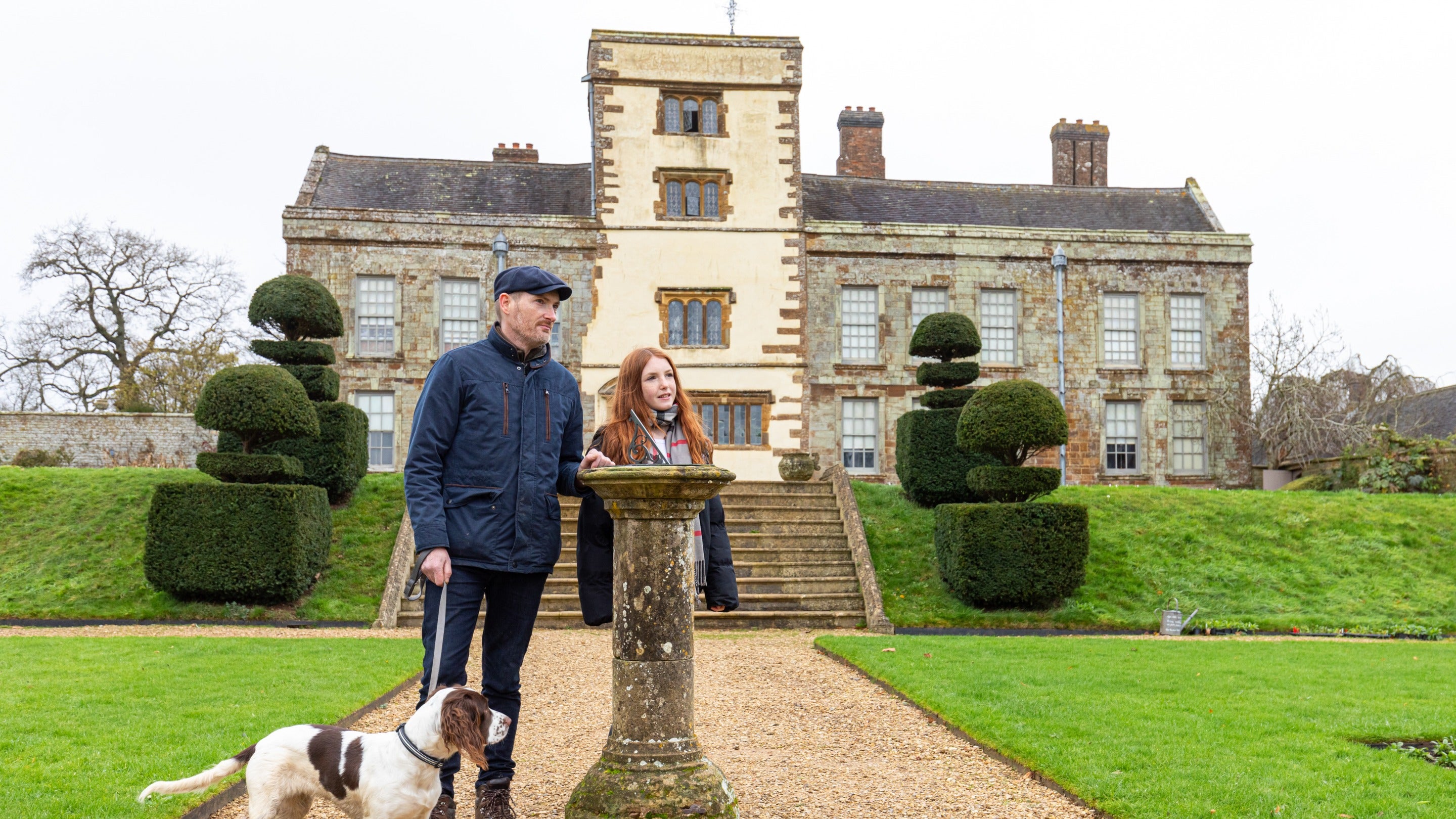 A man and young girl stand in the gardens at Canons Ashby. They are near a sundial with the house in the background. They have a dog on a lead.