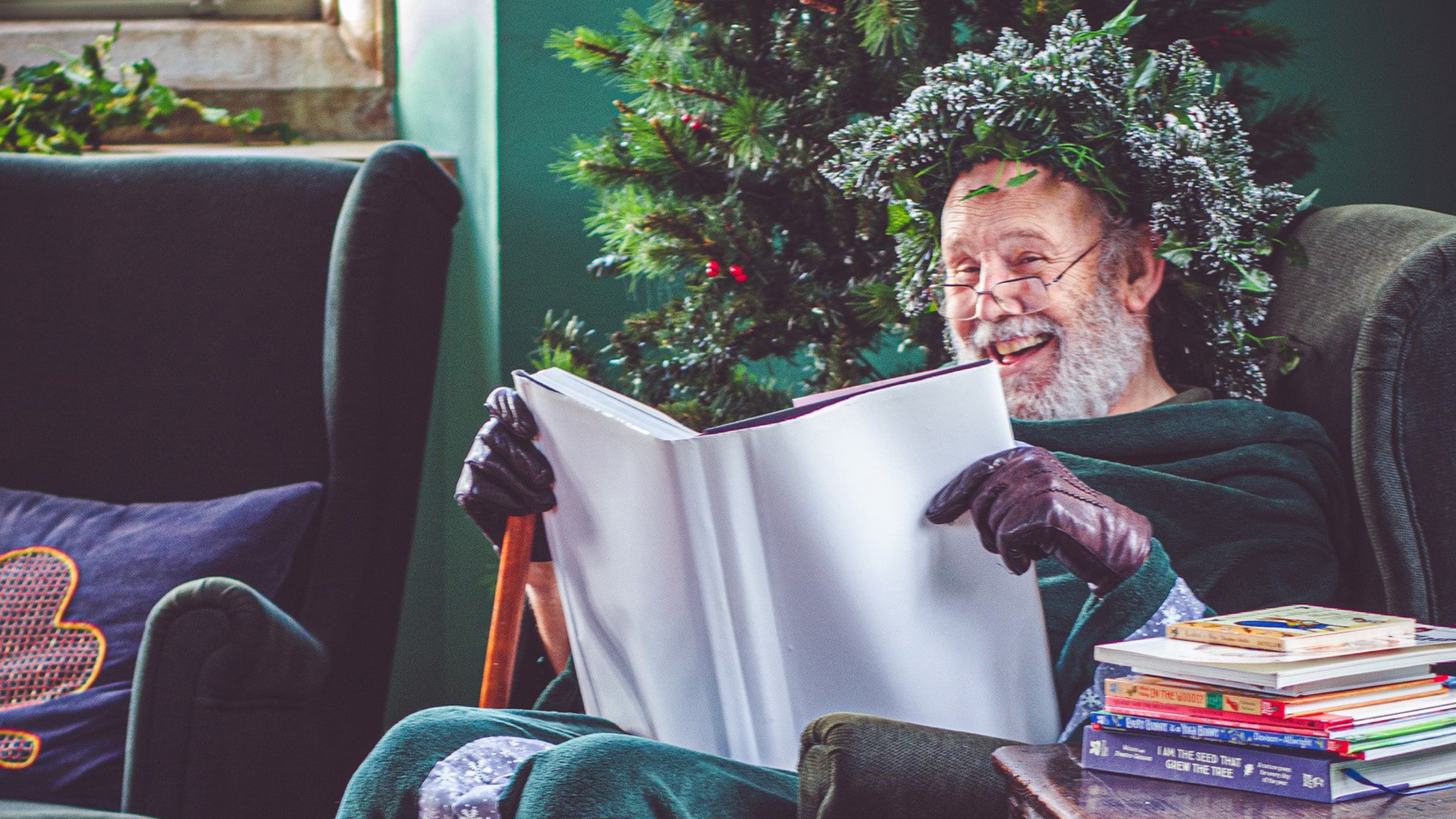 Green Father Christmas reads a story book while sitting in an armchair next to the Christmas Tree.