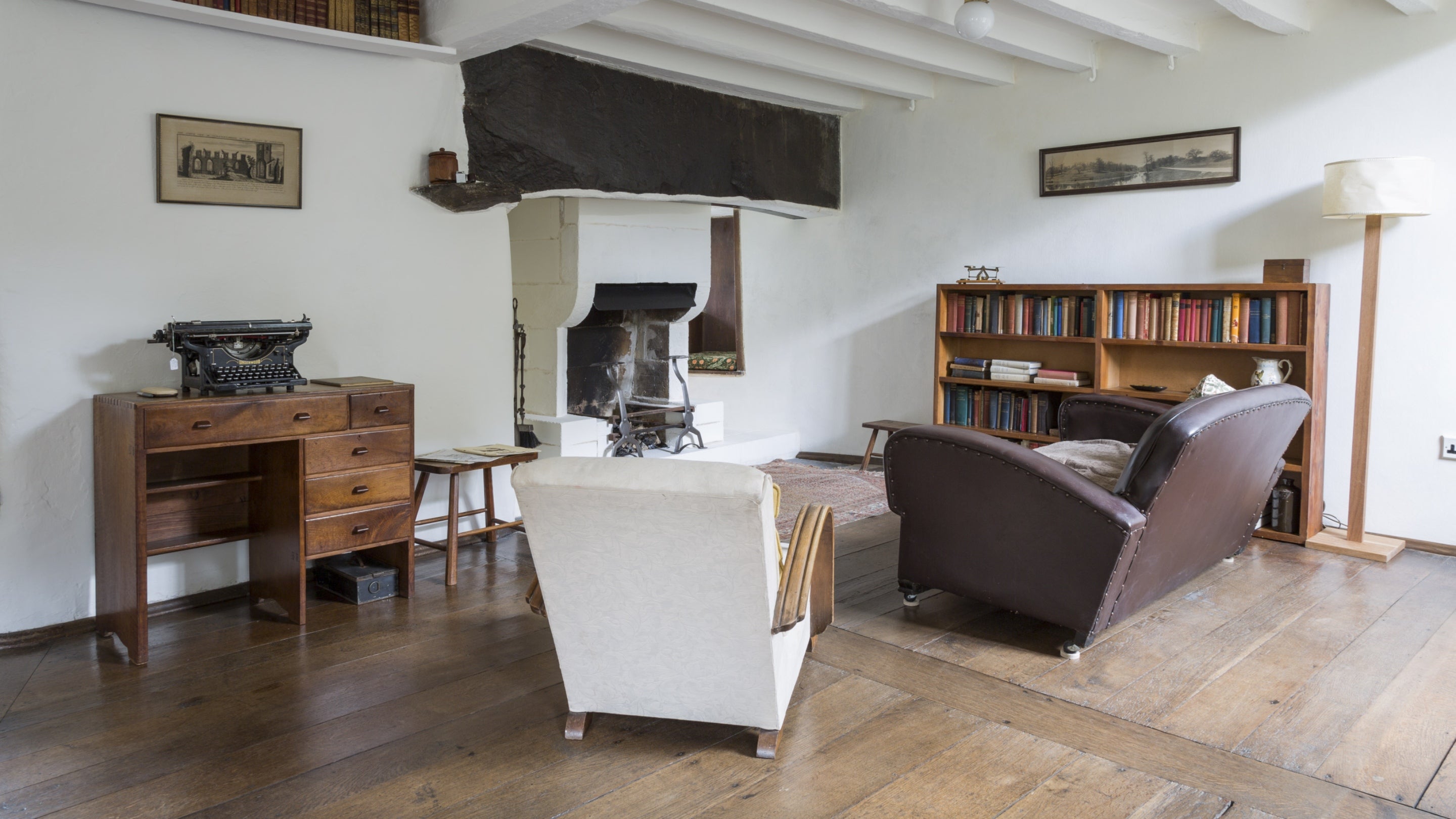 The Sitting Room with white walls and wooden floors at Stoneywell, with a chair and couch surrounding the large fireplace, and bookshelves, a desk and typewriter visible