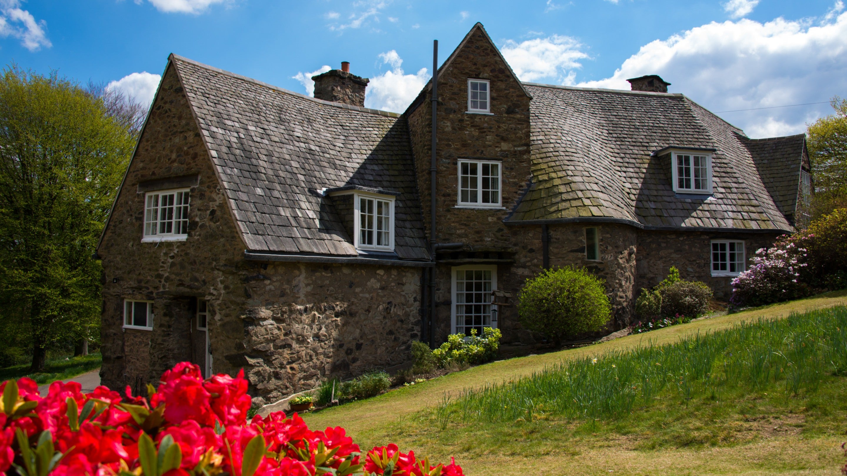A view of the cottage at Stoneywell on a sunny day with red flowers blooming in the foreground and blue skies above