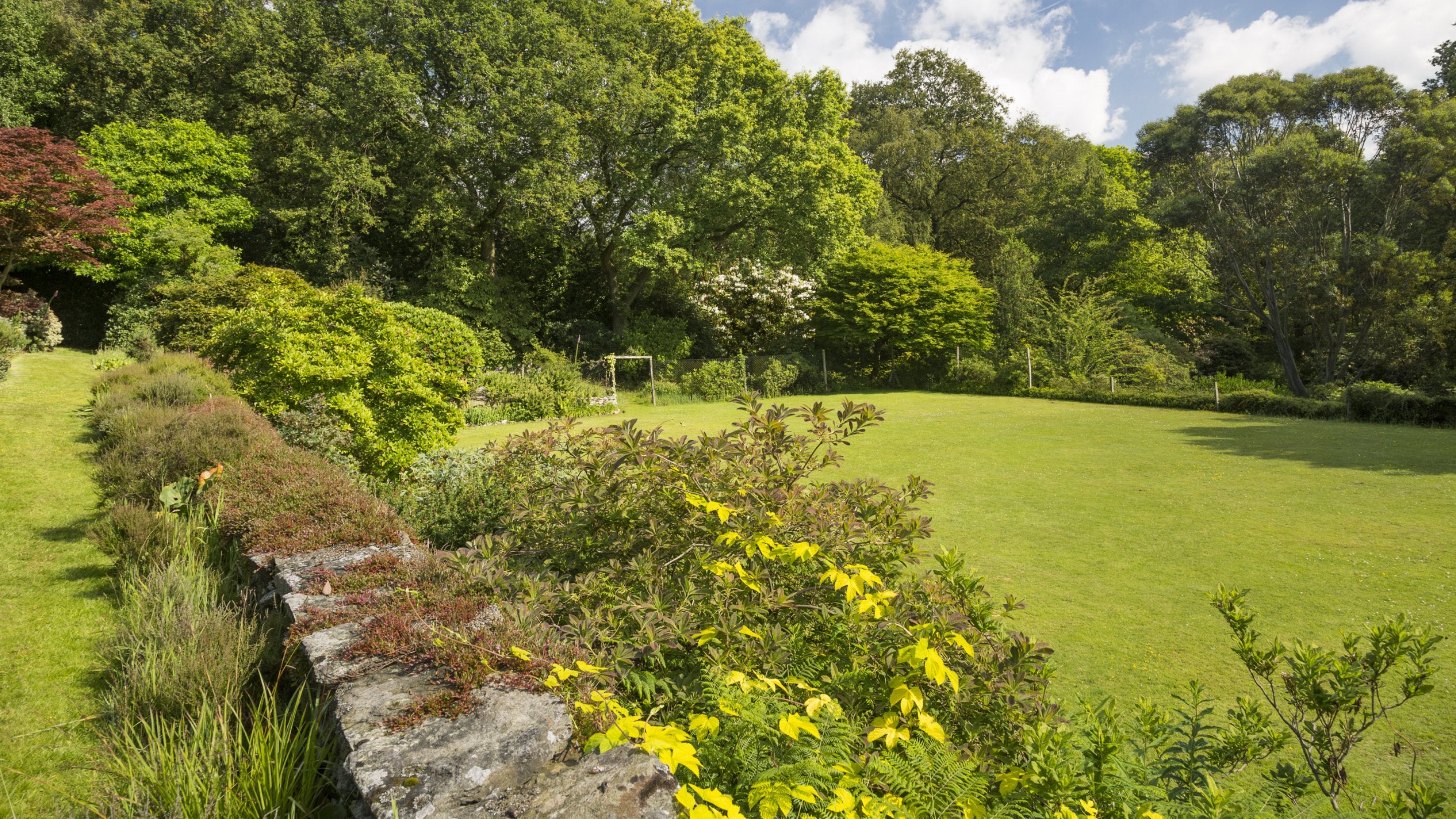 A view over the neatly kept tennis court on a sunny day at Stoneywell with a stone wall and flower borders to its left, and many green trees behind