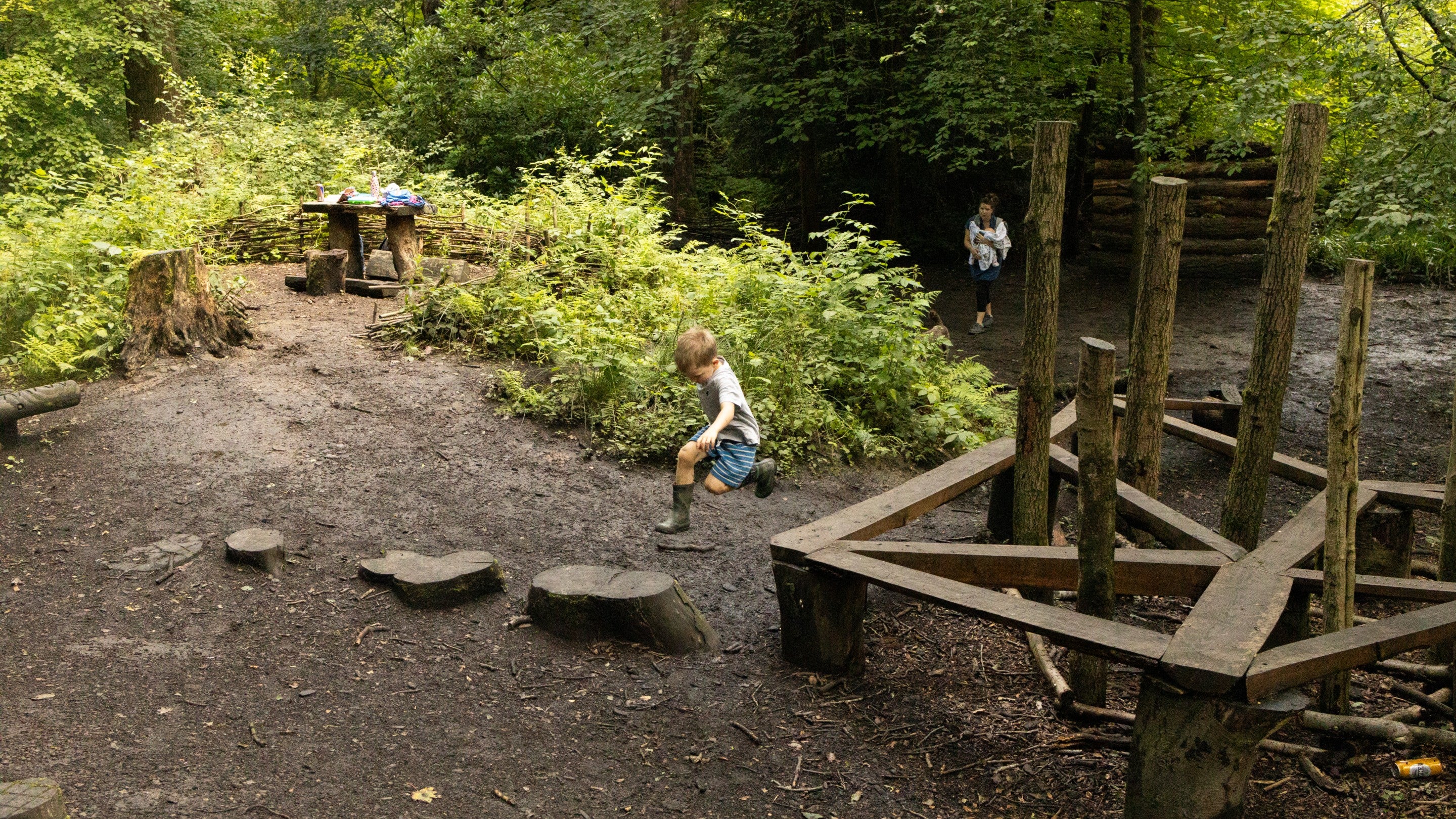 A child in shorts jumping off a wooden structure onto a wooden stepping stone inside a woodland.