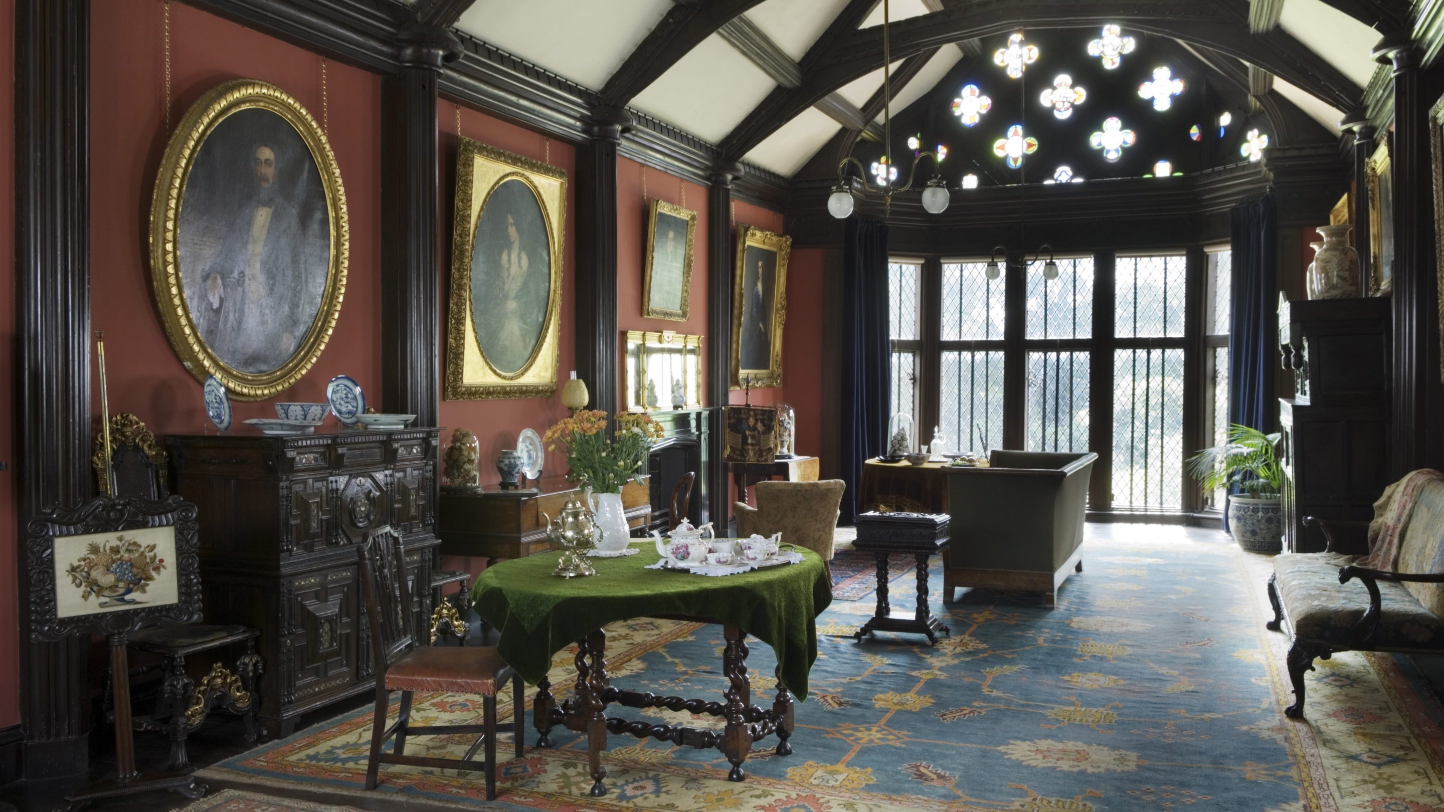 Inside the Drawing Room at Rufford Old Hall, Lancashire, which has exposed beams on the ceiling and is richly furnished with decorative wooden furniture and gold-framed portraits on the walls.