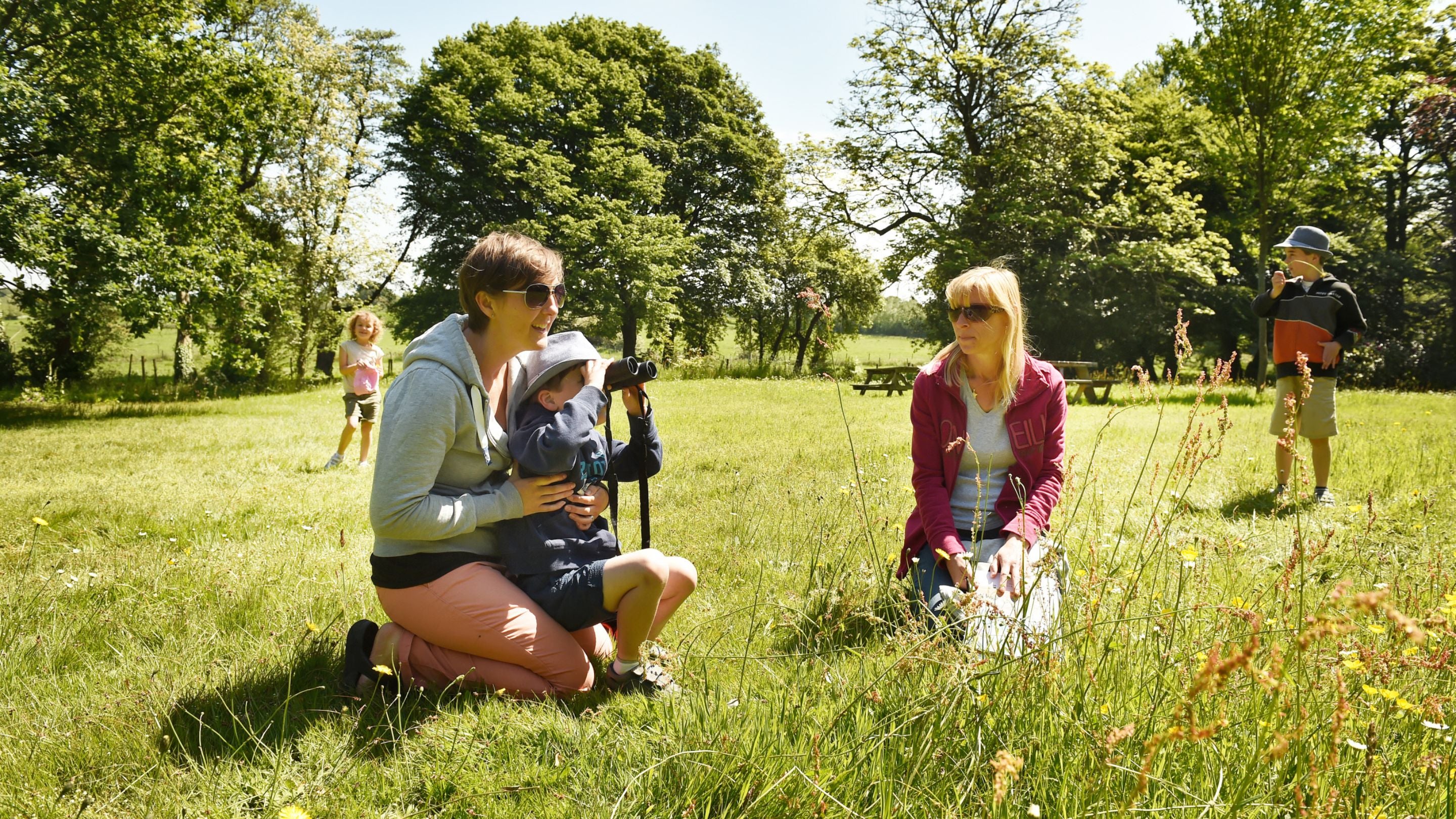 Two adults and three children exploring a grassy meadow, looking through binoculars