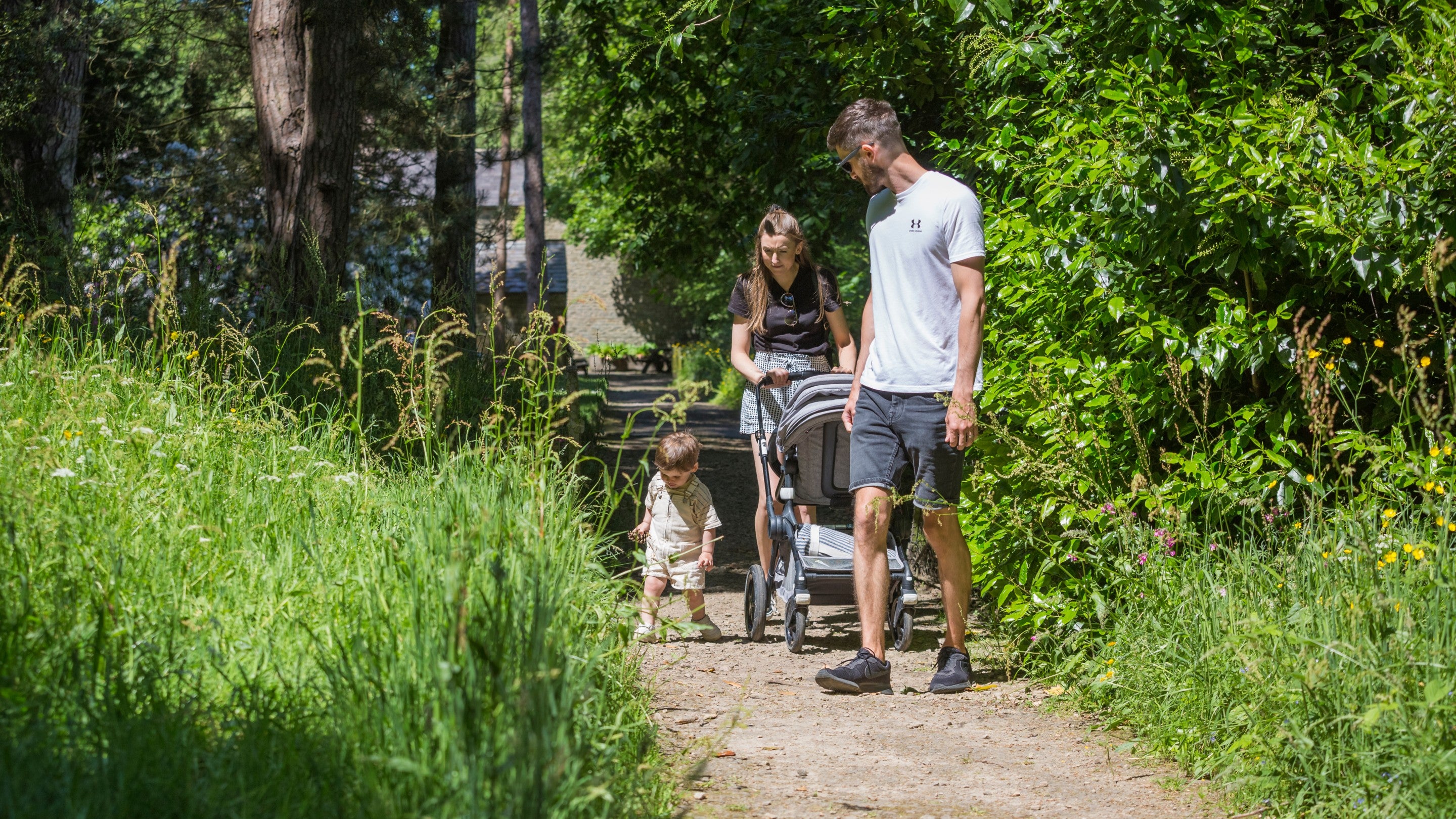 A young couple with a toddler pushing a pushchair down a garden path surrounded by trees.