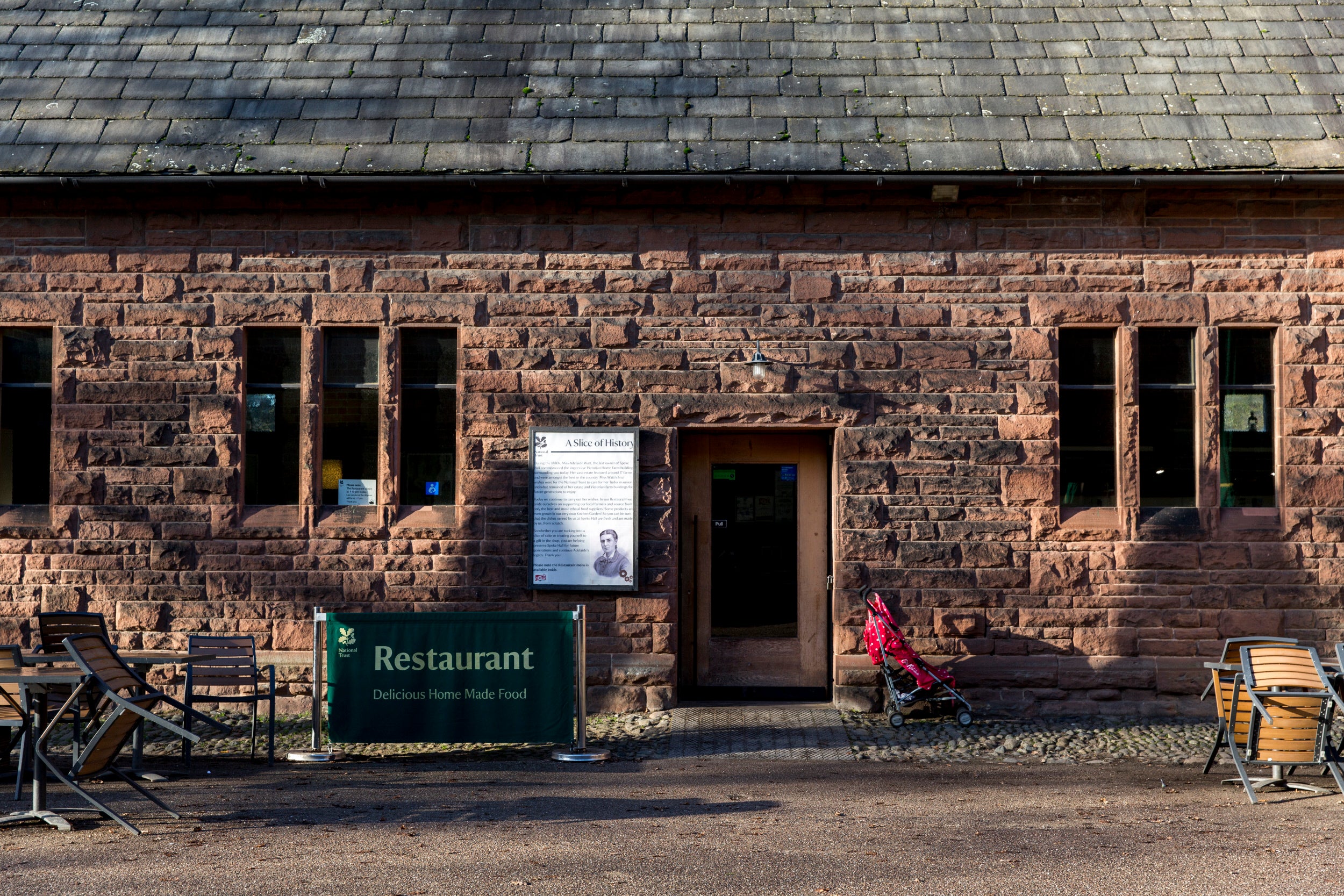 Exterior view of the brick restaurant building, featuring a sign announcing "Restaurant. Delicious Home Made Food," a historical plaque, dark windows, a doorway and a child's stroller. Outdoor seating is situated nearby.