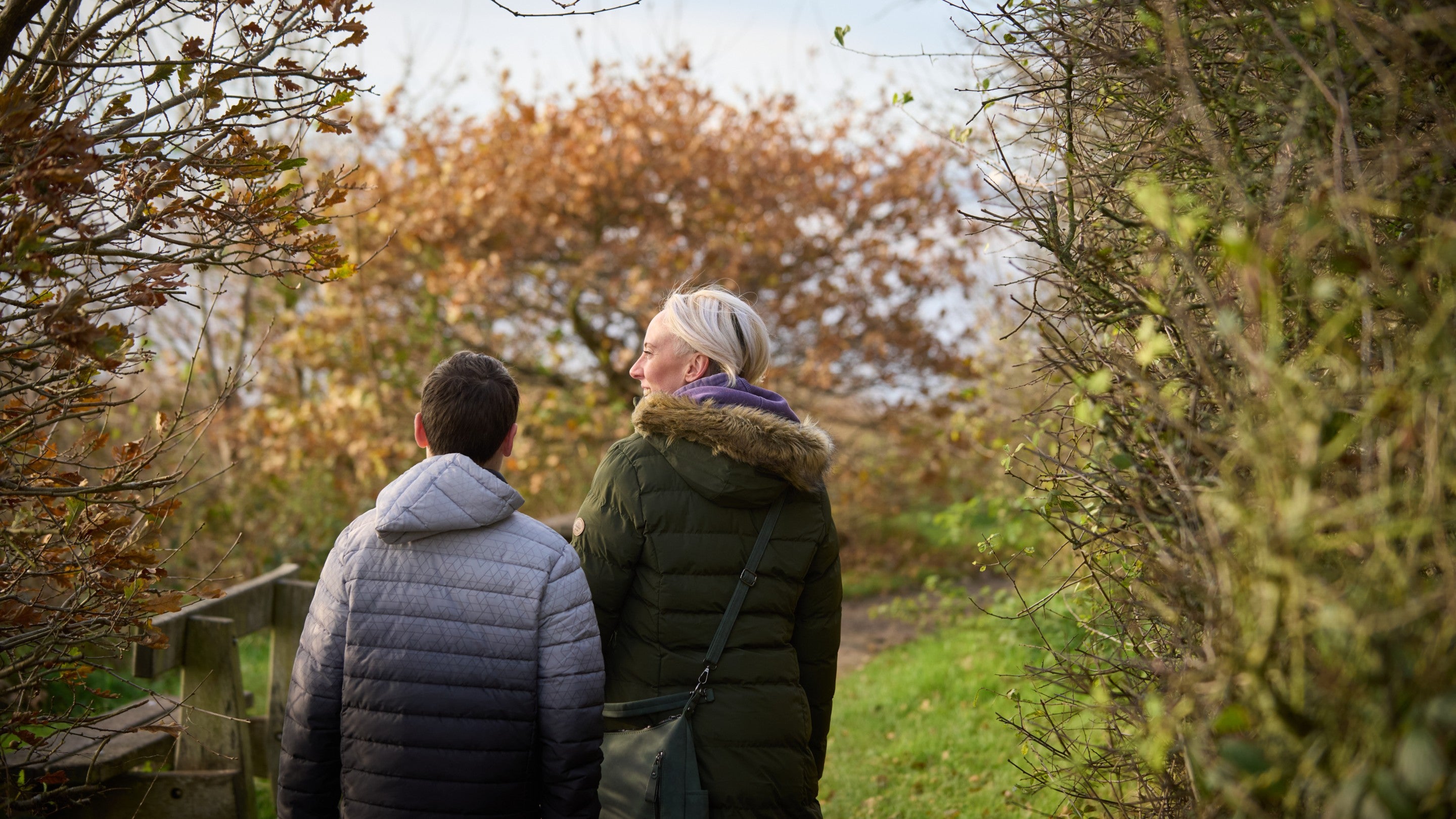 Two people walking down a narrow path framed by dense shrubs and trees, with autumn leaves visible ahead and water in the distance.