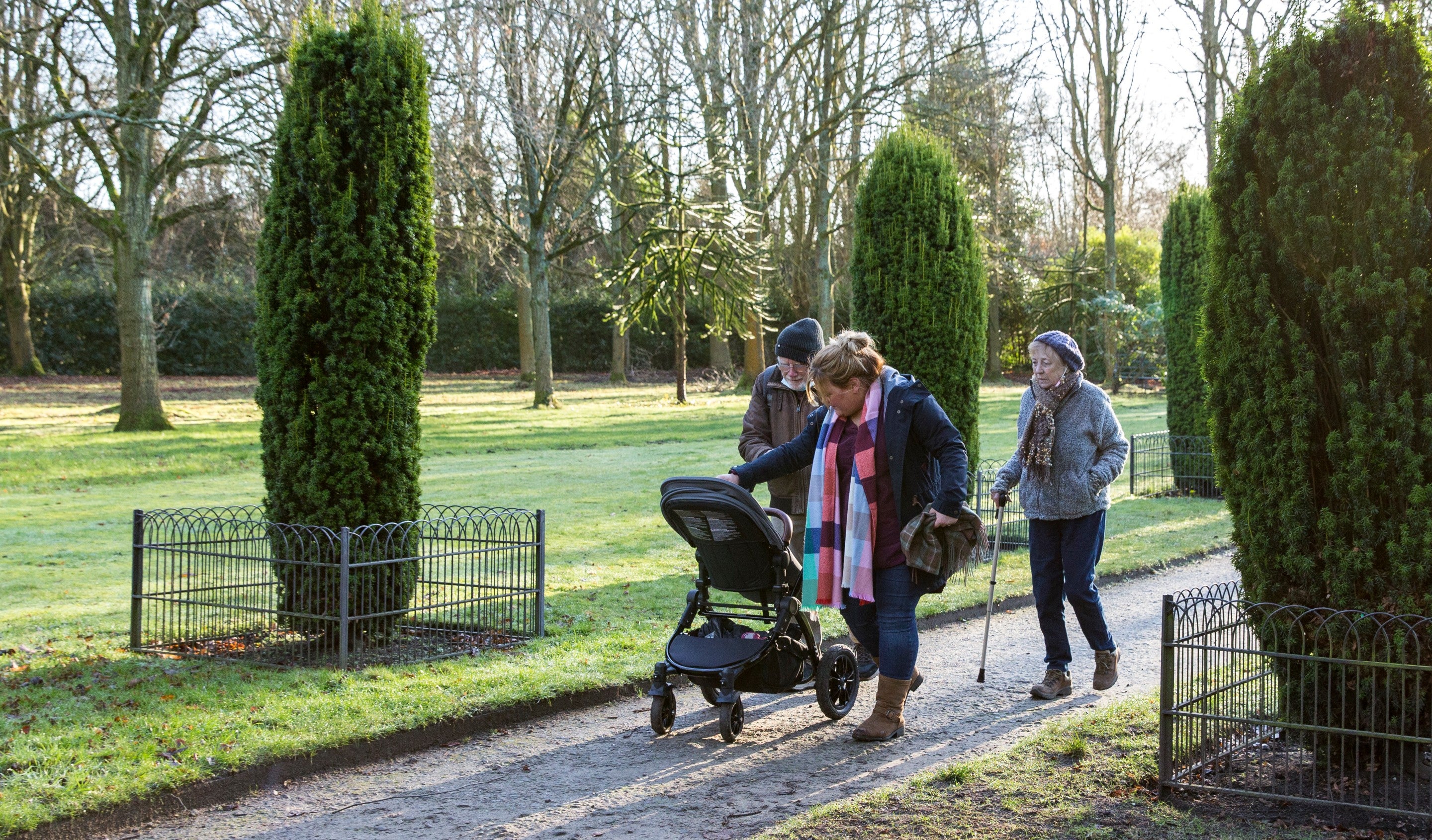 A multi-generational family of three adults and one child walk through the gardens at Speke Hall. They are dressed for winter and pushing a pram.
