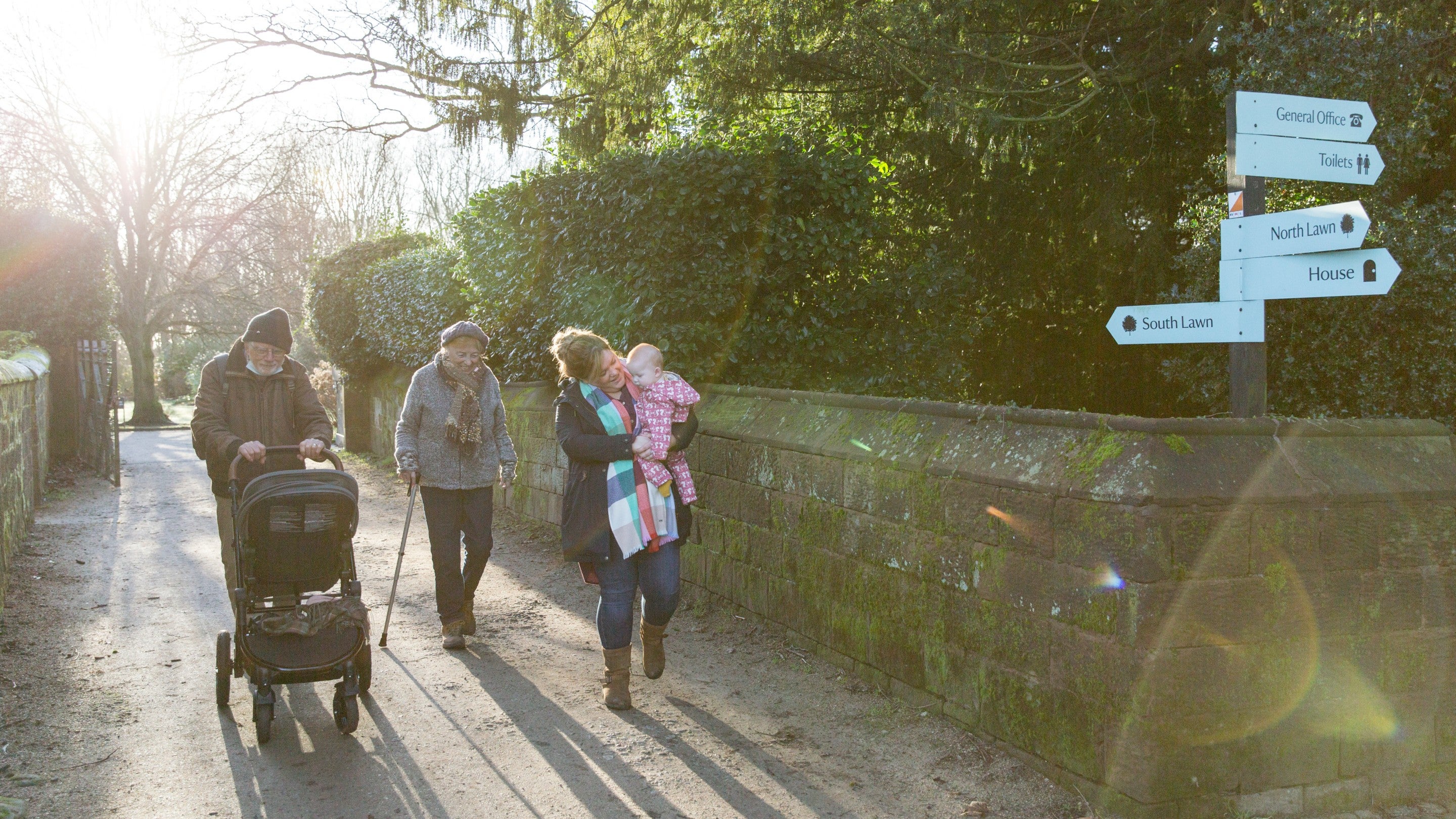 A multi-generational family of three adults and one small child walking on a path at Speke Hall. They are dressed for winter and the man is pushing a pram.