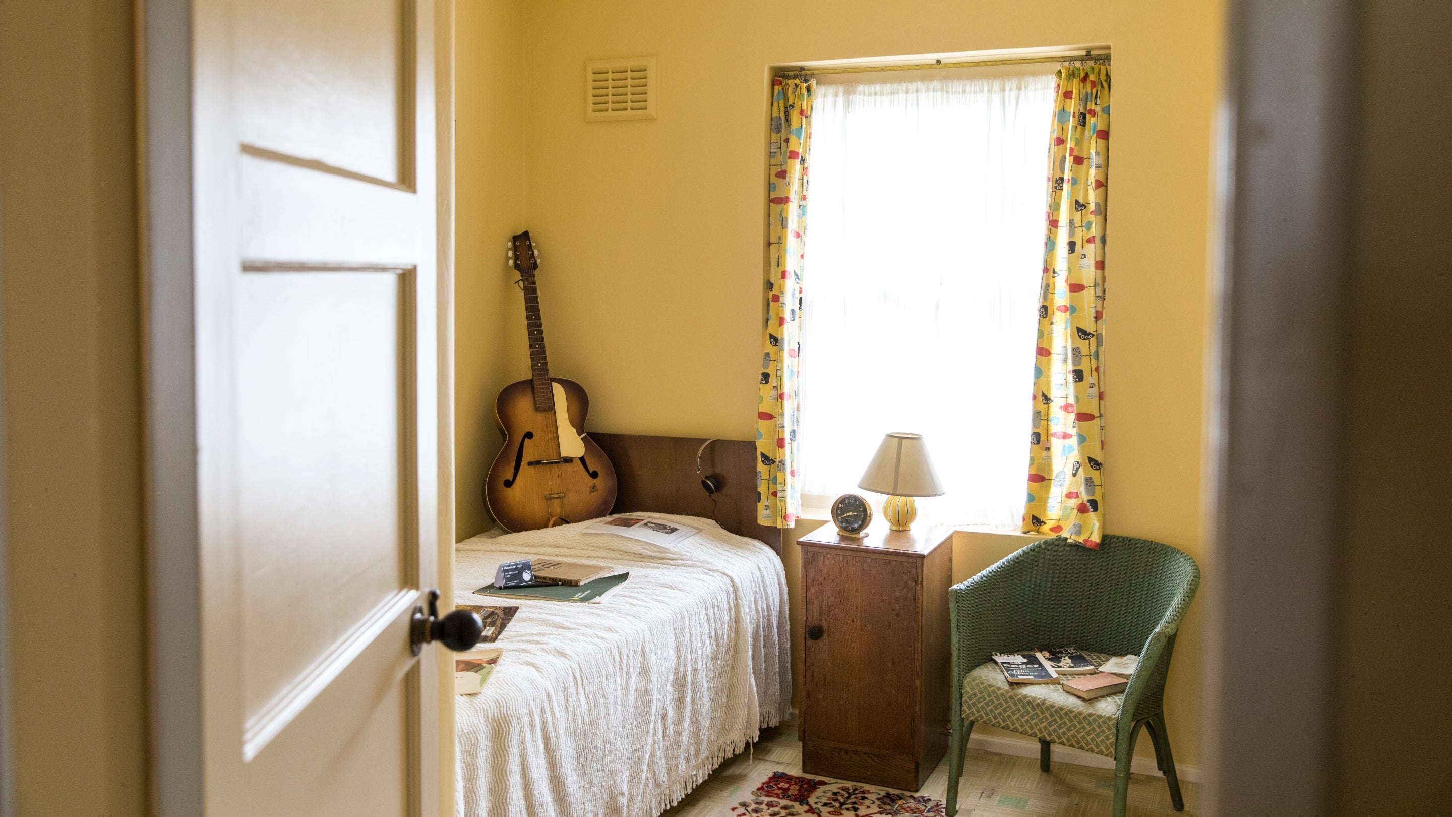 A view from the doorway of Paul McCartney's childhood bed, guitar and green chair in a small cream-coloured room.