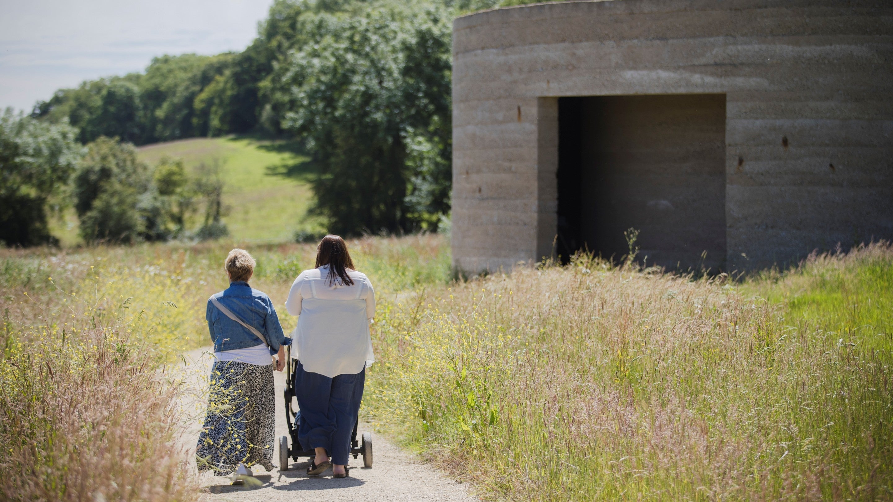 Two adults walk along a narrow path through tall grass, pushing a stroller. To the right stands a round concrete architectural artwork structure with a large rectangular opening. Rolling countryside and trees extend into the distance under a bright sky.