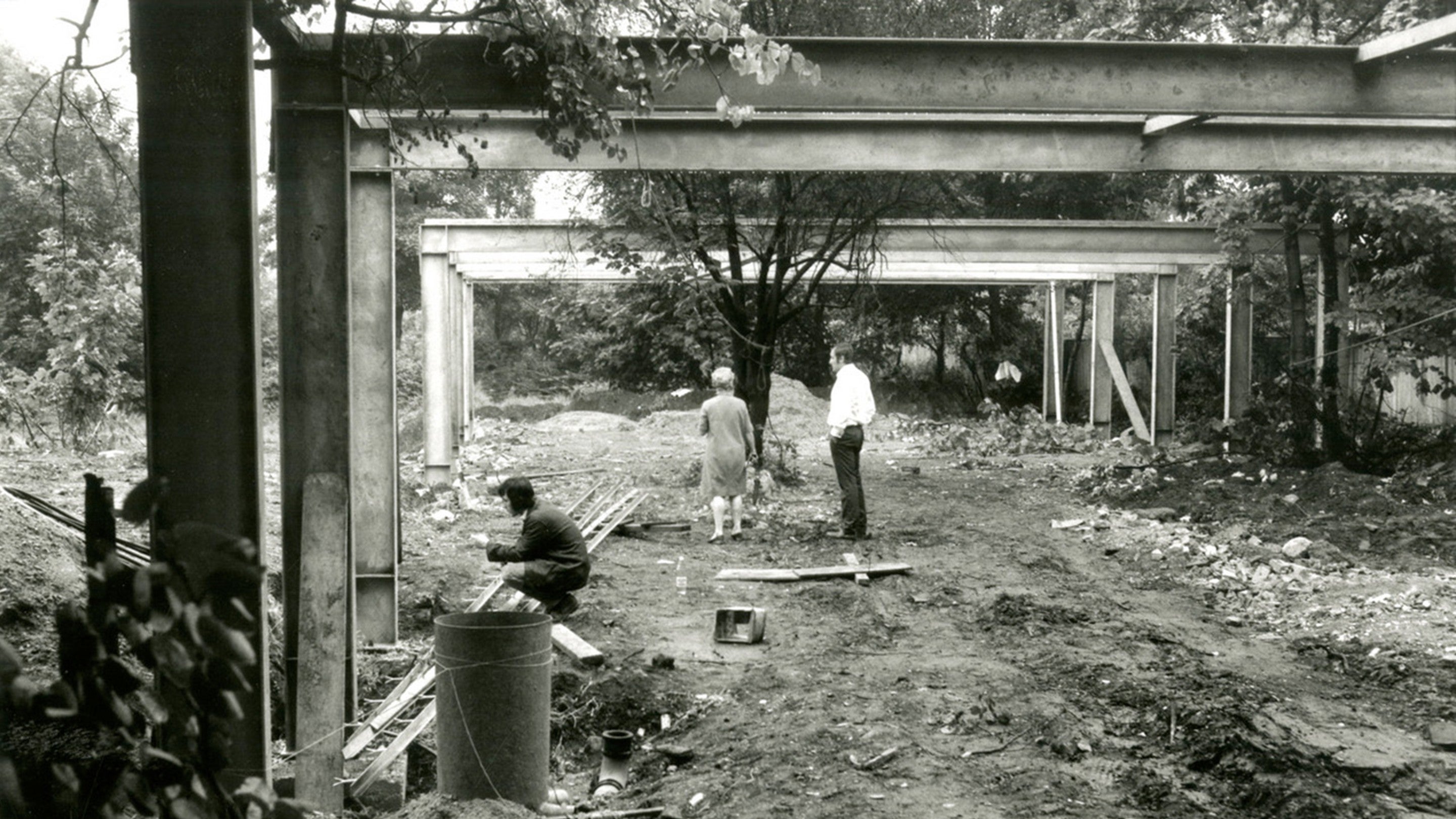 A black and white photo of a building site. Three people access the site. Steele beams form the skeletal shape of a single storey