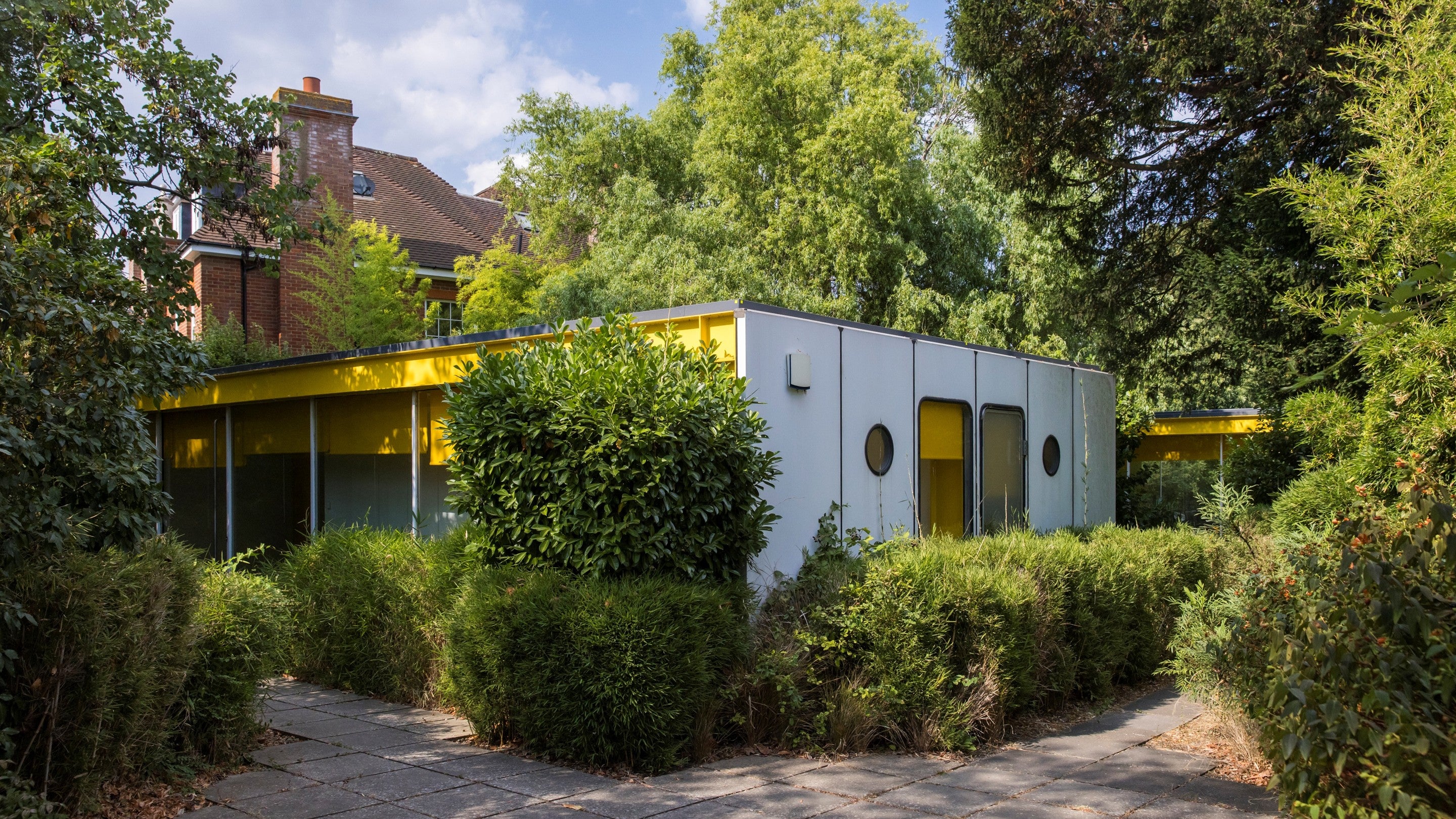 A modern bungalow with a block white and yellow walls and full height windows. Trees and hedges in the foreground.
