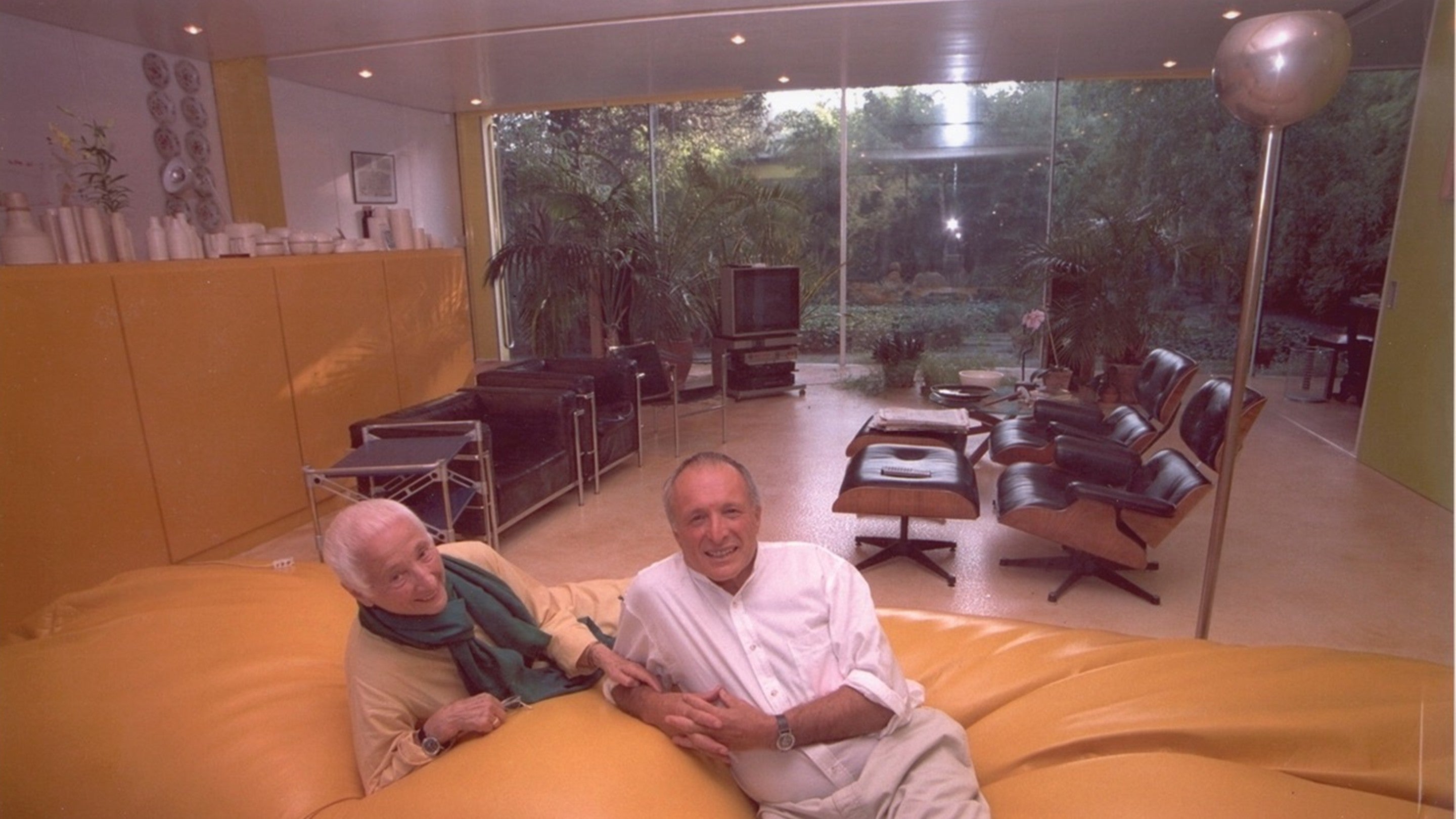 Richard Rogers, co-designer of 22 Parkside and his mother, Dada Rogers lounge on a giant yellow bean bag in the foreground of the photo. A modern living room with four black arm chairs and yellow cabinets are in the background.