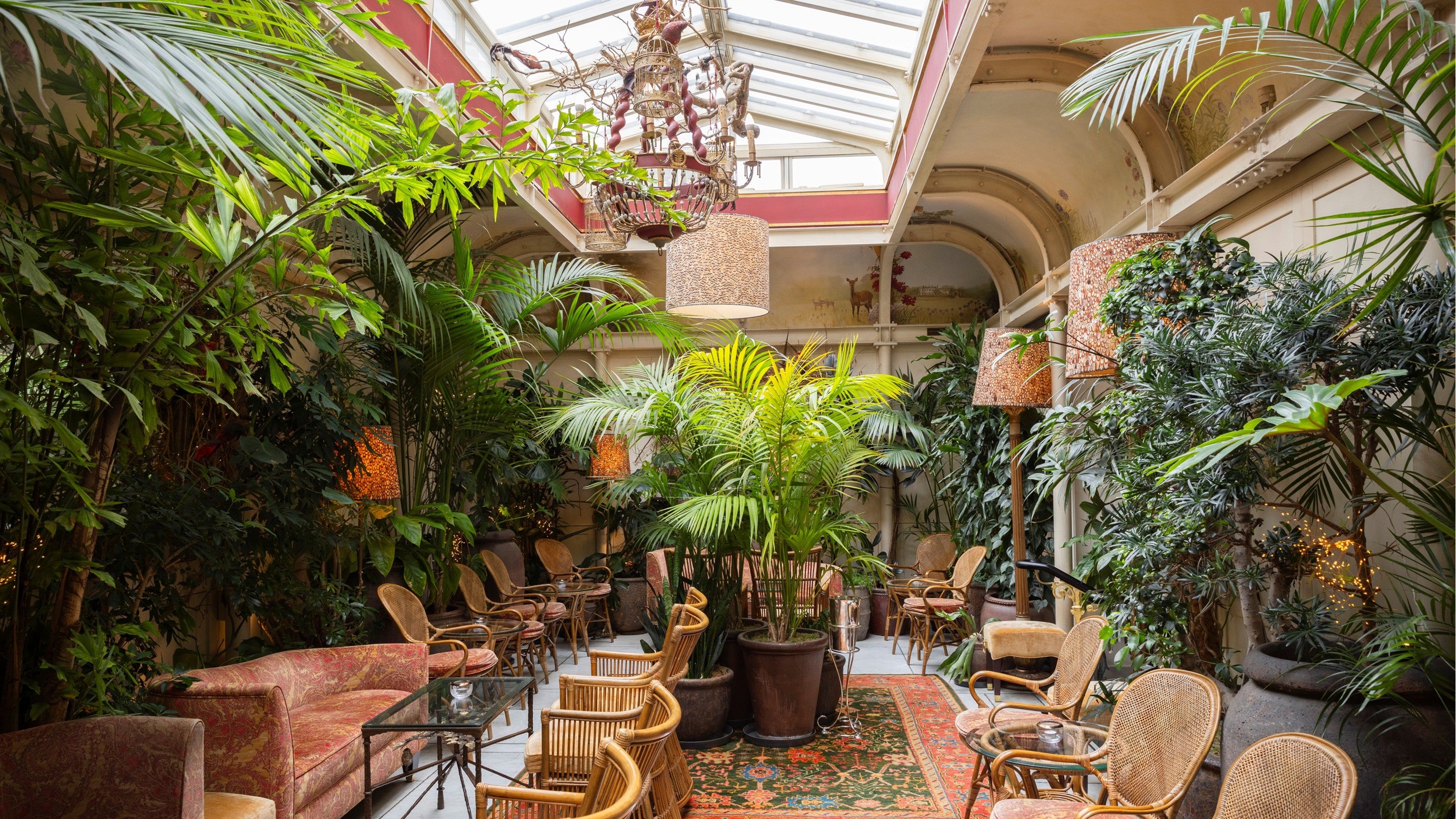 Light-filled winter garden with a glass roof, abundant tropical plants, wicker chairs, patterned rugs, and intimate seating areas arranged among large potted palms and greenery.
