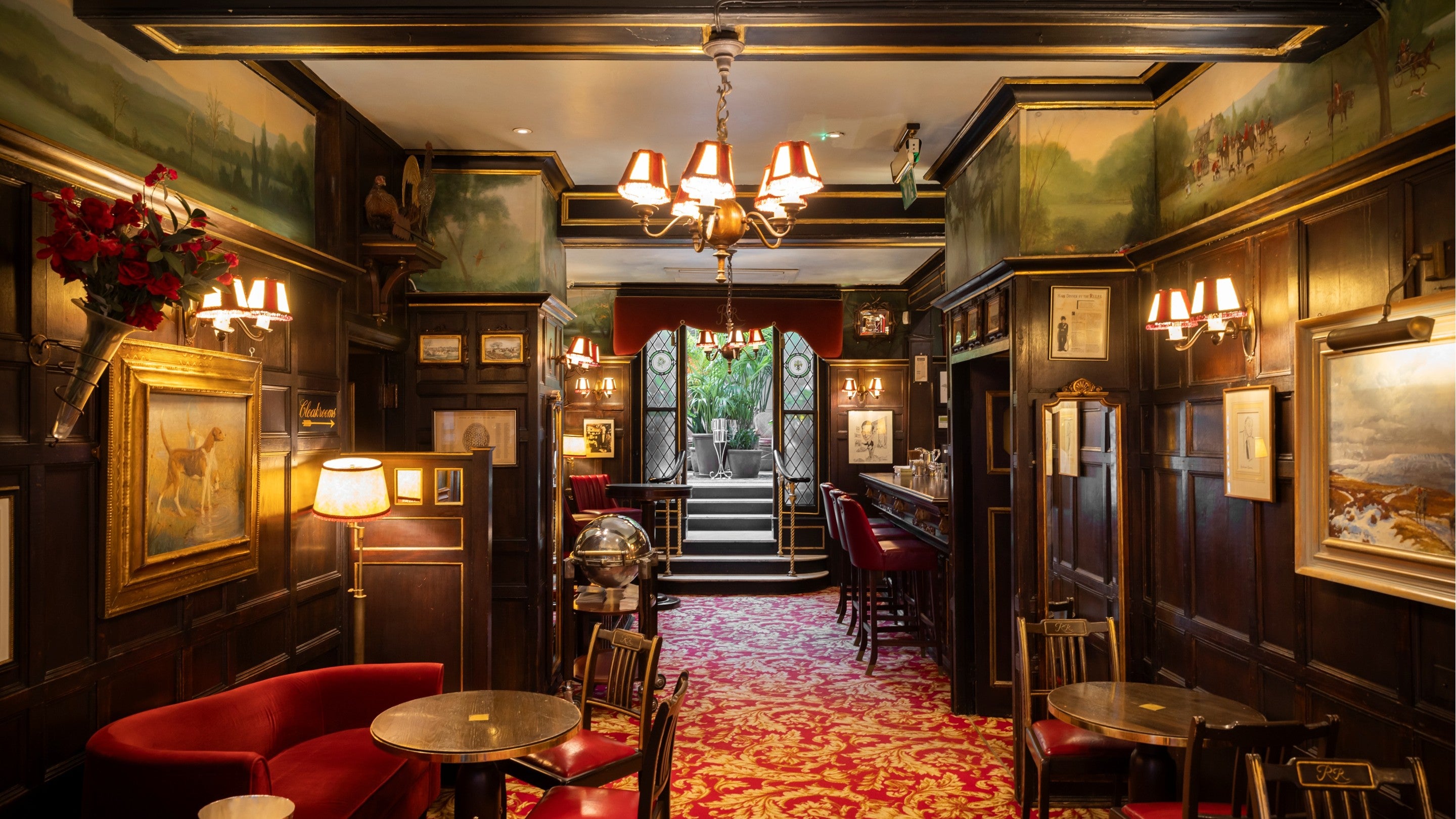 View through an elegant cocktail bar with dark wood panelling, red upholstered seating, framed artwork, and ornate lighting, leading towards a bright glass-roofed winter garden filled with greenery.