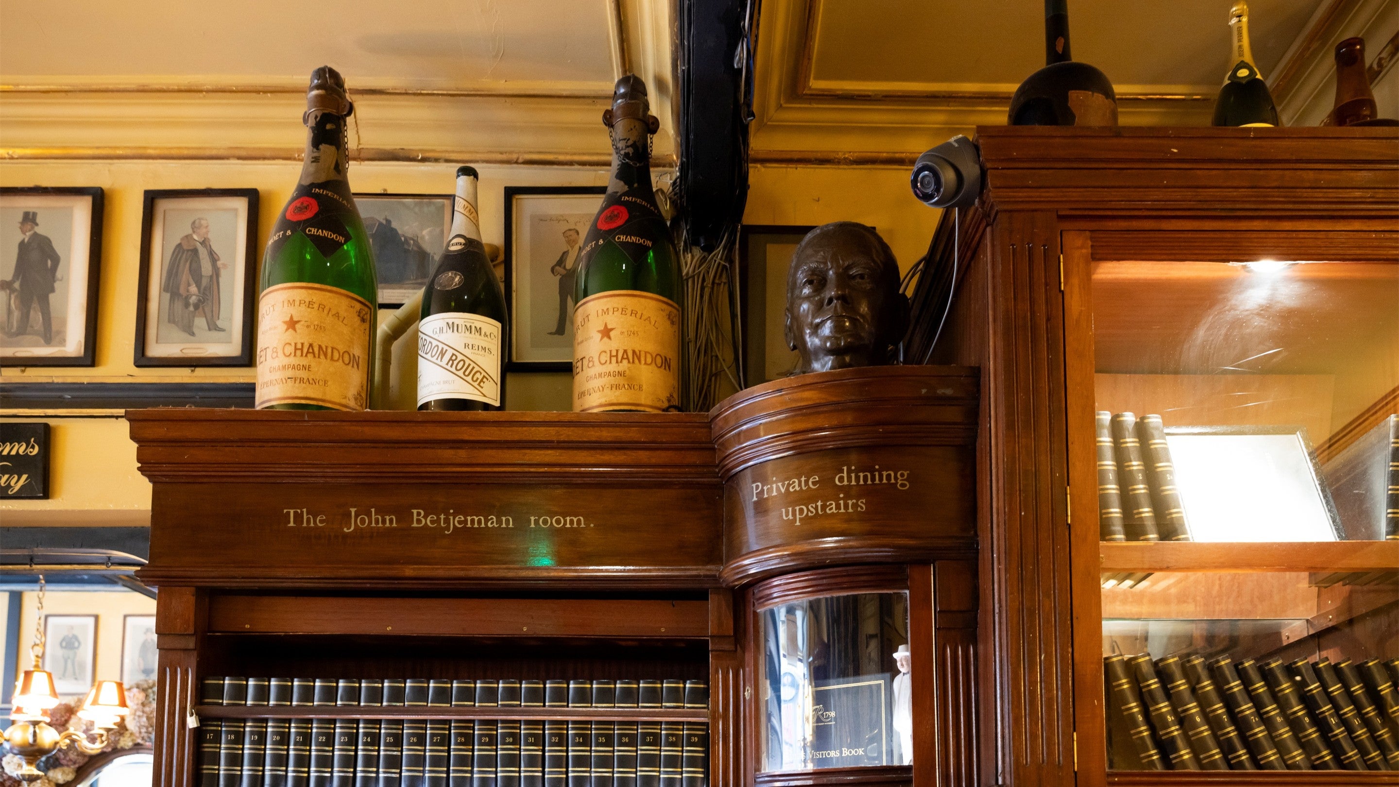 Historic wooden cabinet lined with leather-bound visitor books, featuring engraved signs for the John Betjeman Room and private dining area, with oversized champagne bottles, framed illustrations, and a bronze bust displayed above.
