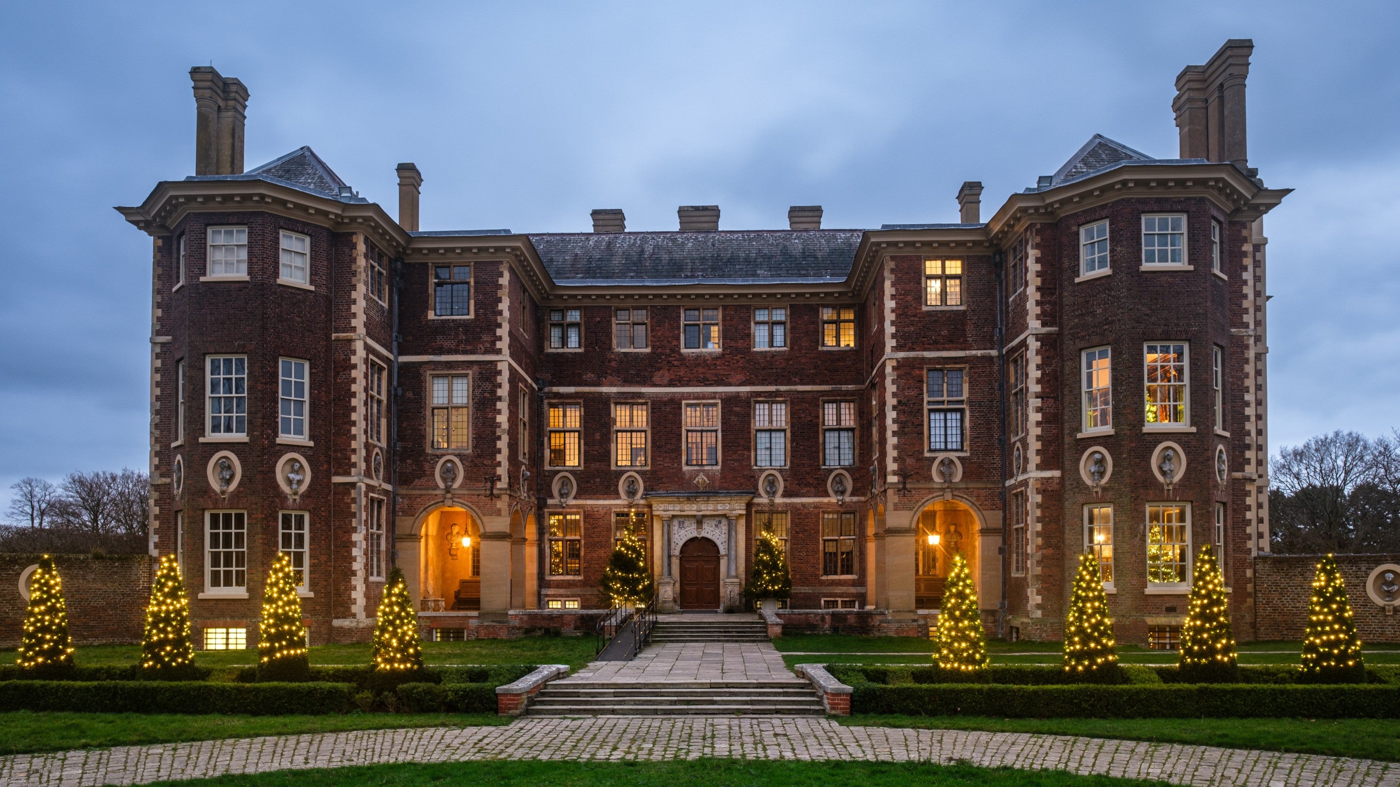 Christmas trees and lights outside Ham House, Surrey with grass and a path in the foreground and a cloudy sky above