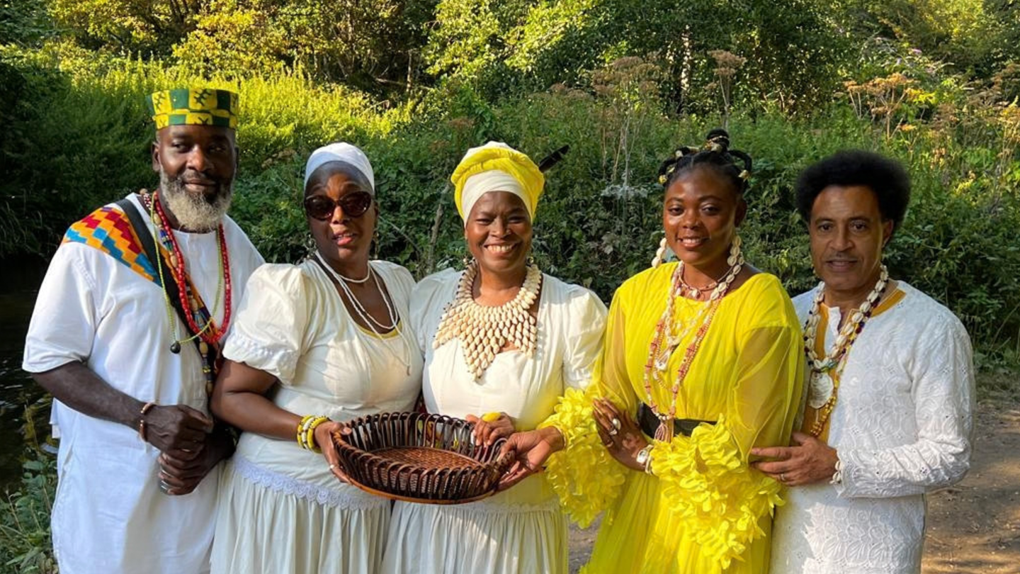 A group photo of people dressed in traditional African clothing in a outdoor setting at Morden Hall Park with trees and shrubs in the background.