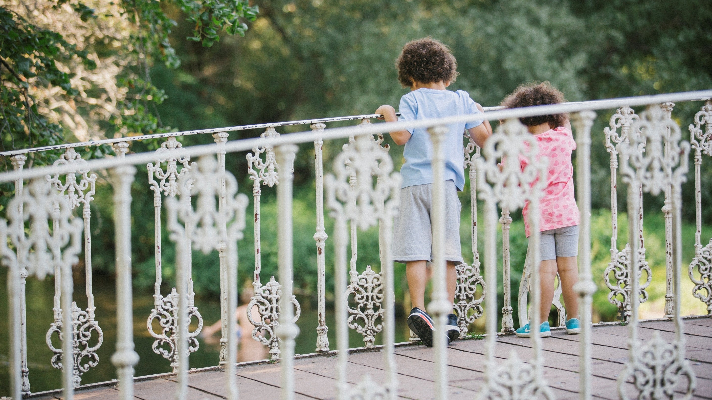 Two young children walk across an ornate white metal bridge with decorative railings, looking over the side toward water below. Trees and greenery fill the background.