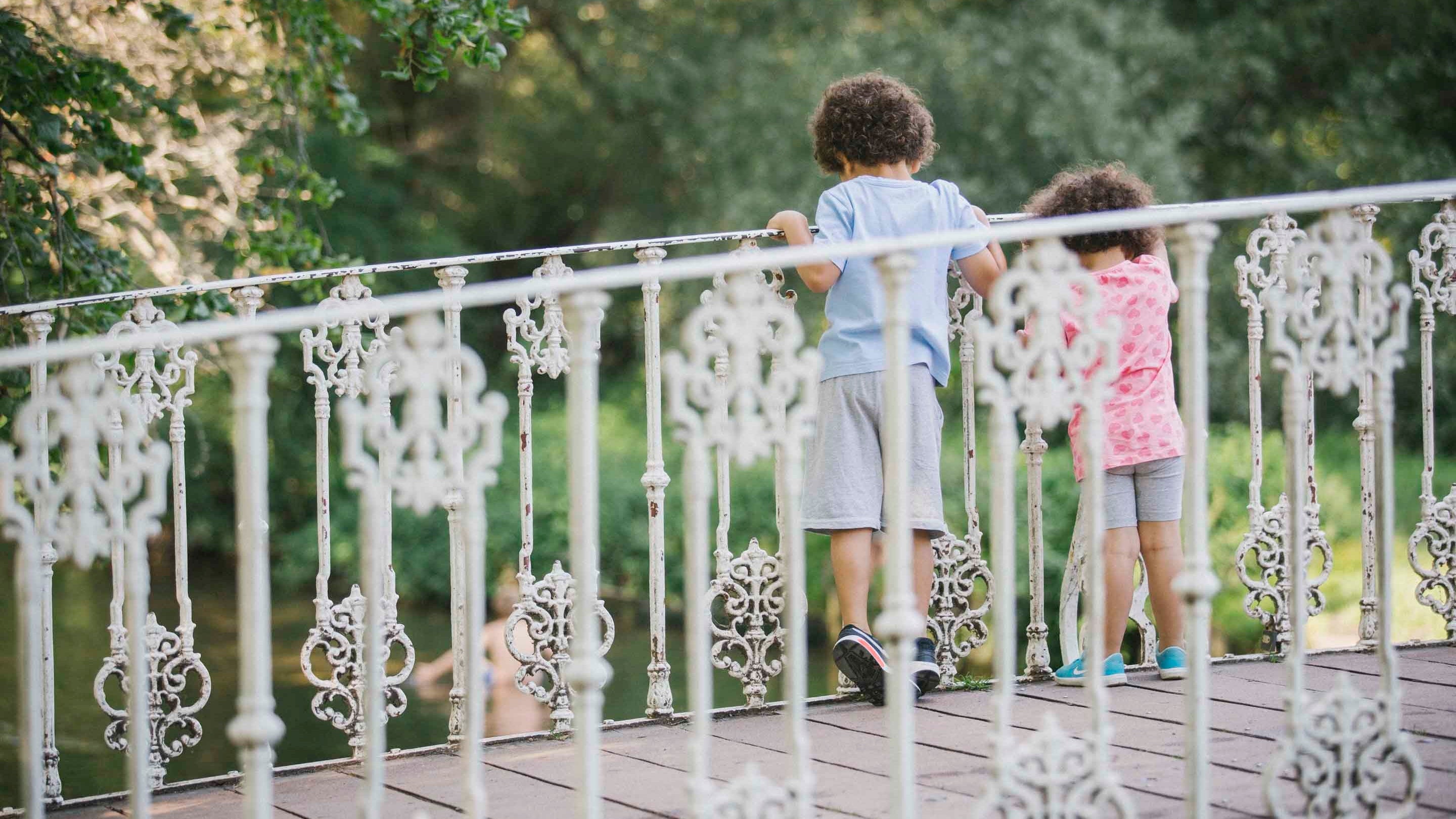 Two children stand on the white bridge over the River Wandle at Morden Hall Park, London. Their backs can be seen as the look over the bridge railings to look in the water below.