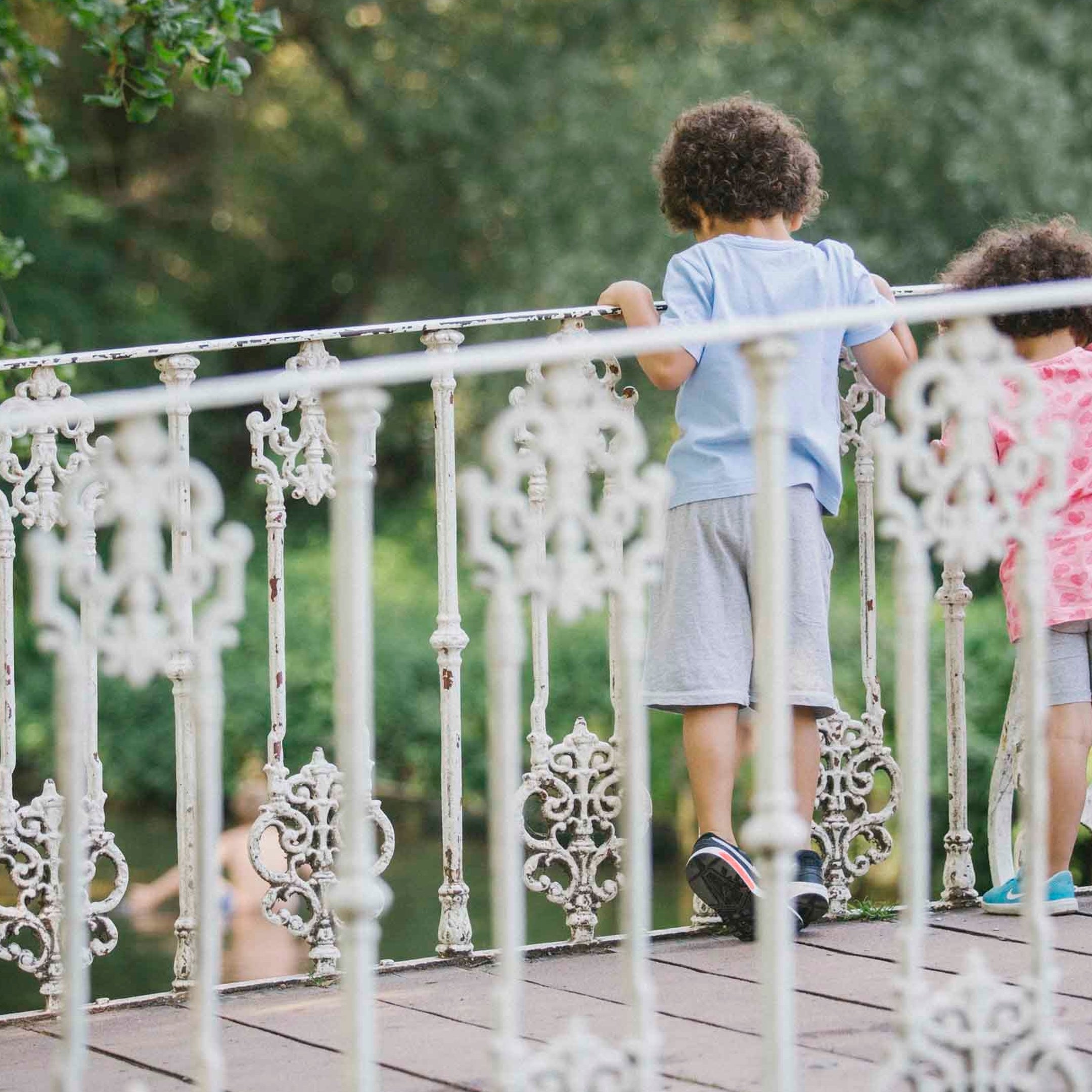 Two children stand on the white bridge over the River Wandle at Morden Hall Park, London. Their backs can be seen as the look over the bridge railings to look in the water below.