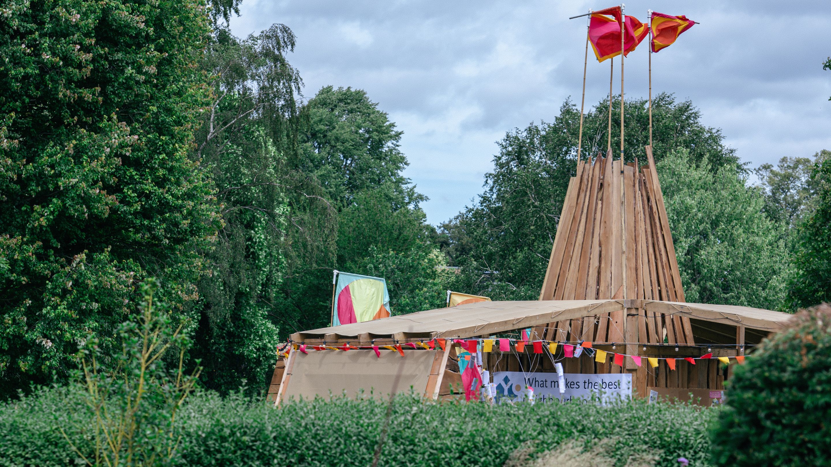 A wooden structure in a green woodland setting seen from behind some bushes with blue sky in the background.  The structure is adorned with bunting with large flags flying above. The structure is part of the Let Hope Blossom public artwork.