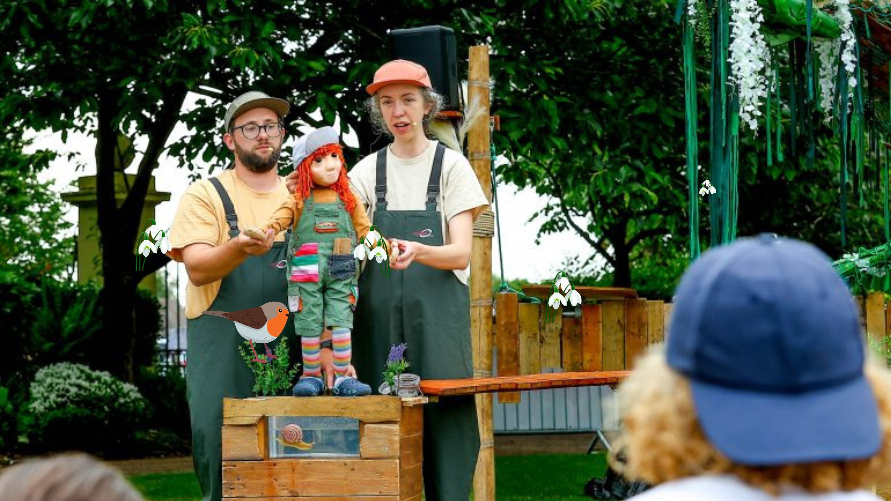 A photography of two puppeteers manipulating a small puppet on a table in an outside setting with audience watching. They are all wearing green dungarees and a peak cap.