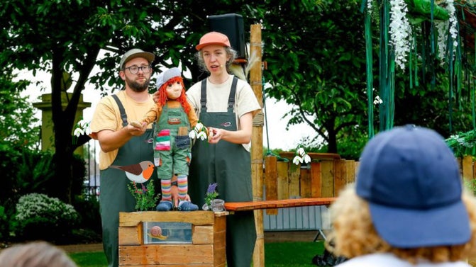 A photography of two puppeteers manipulating a small puppet on a table in an outside setting with audience watching. They are all wearing green dungarees and a peak cap.