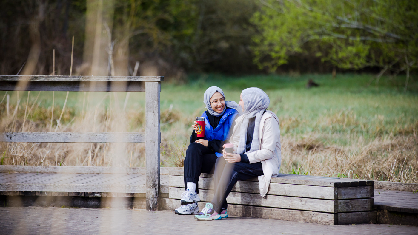 Two women sitting on a bench on the boardwalk at Morden Hall Park, holding reusable coffee cups, smiling and talking