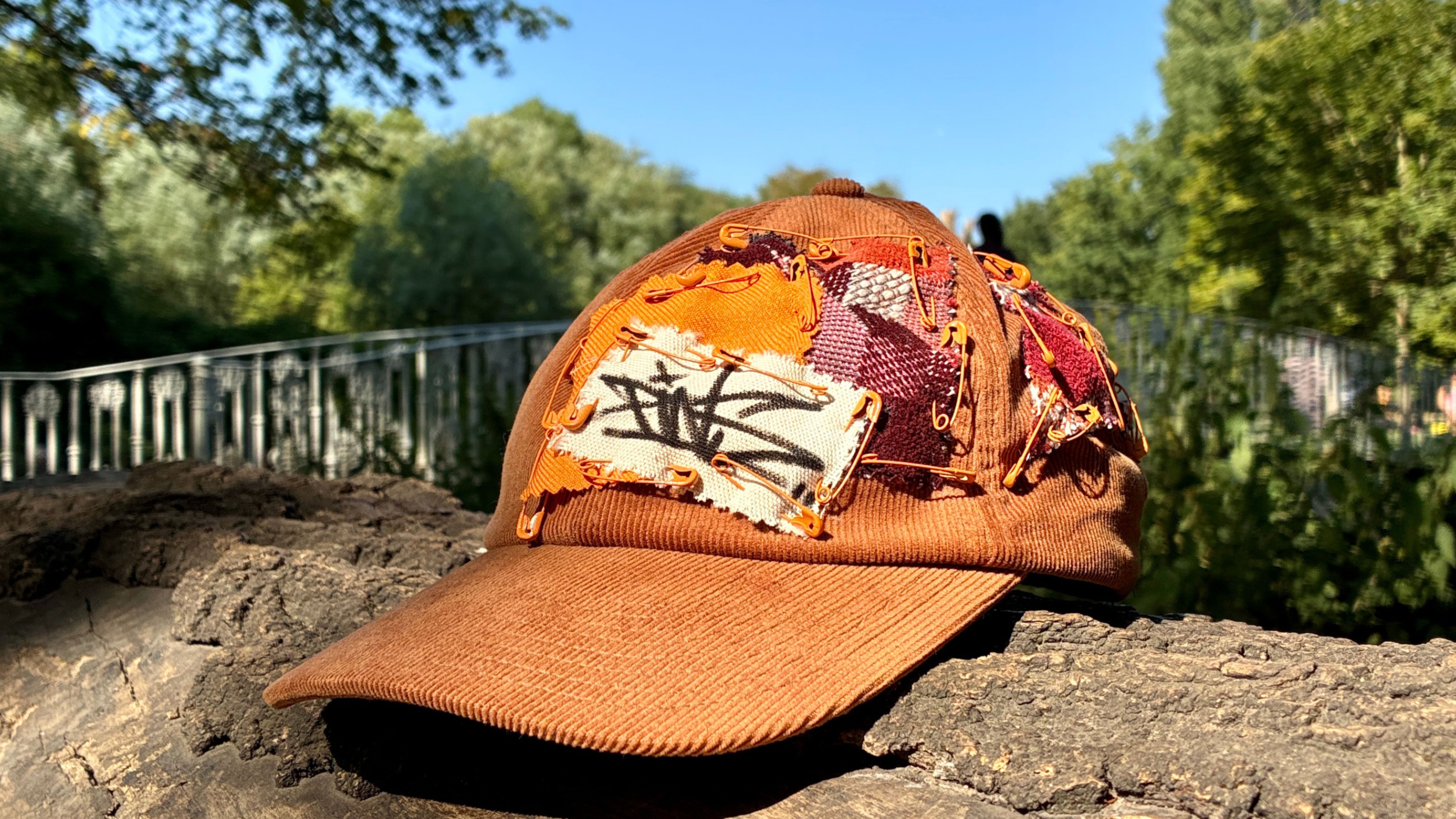 Photography of a brown customised peak cap sitting on a log with the Morden Hall Park white bridge in the background