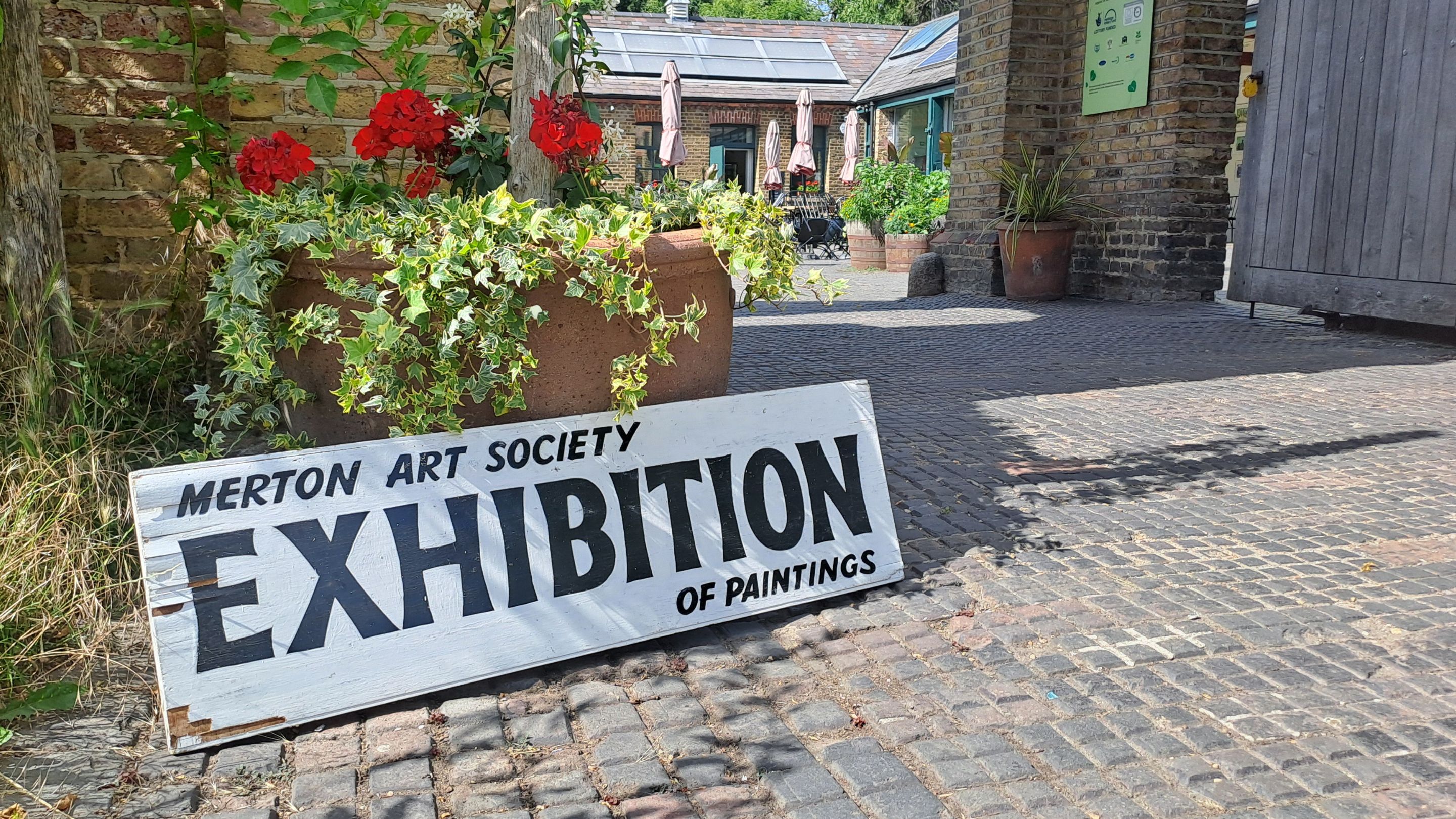 A photography of a wooden board with the text 'Merton Art Society Exhibition of Plainings' leaning against the large plant pot in front of an arched entrance way.