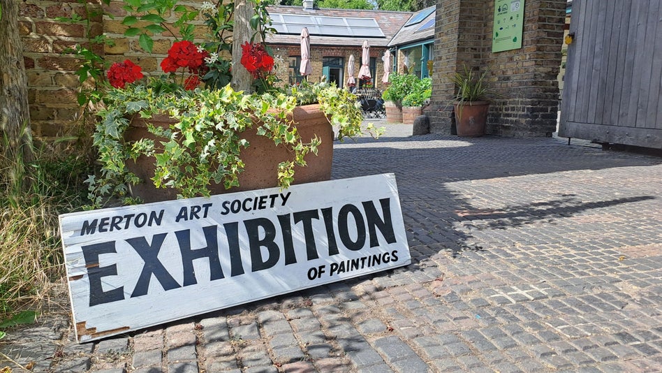 A photography of a wooden board with the text 'Merton Art Society Exhibition of Plainings' leaning against the large plant pot in front of an arched entrance way.