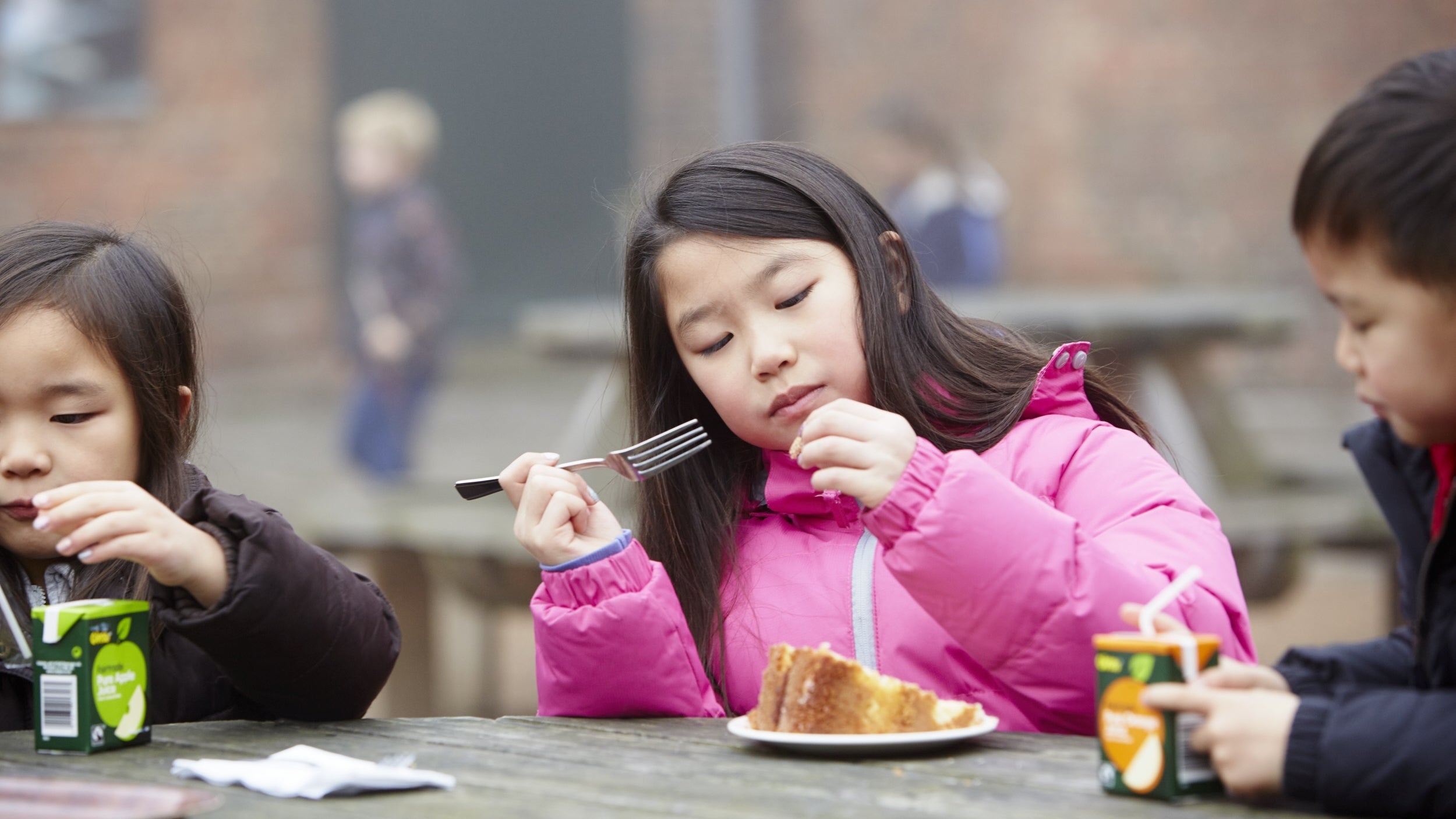 Three children sitting at a table outside while eating cake and drinking juice boxes