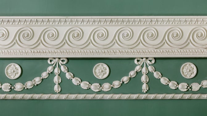 Close-up of ornate white plaster moulding featuring scrolls, floral motifs, and ribbon swags set against a pale green wall.