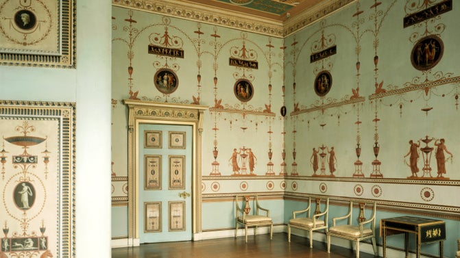Pale blue historic room decorated with intricate red and gold classical motifs, featuring painted medallions, a decorative door, and a row of delicate chairs along the wall.
