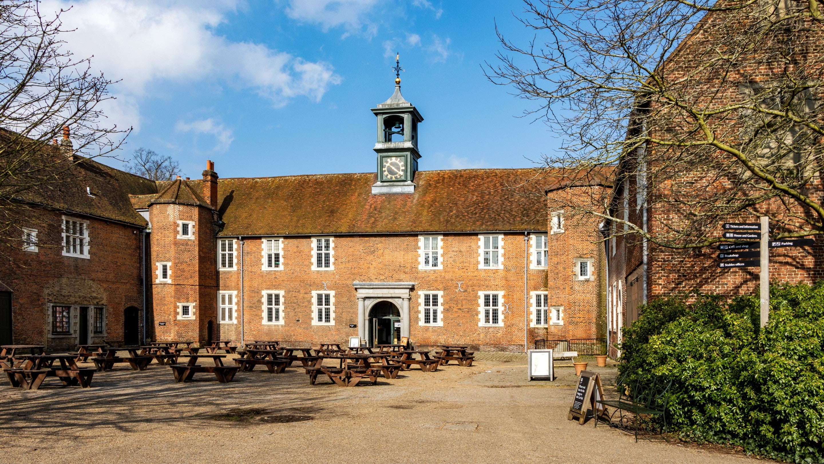 Historic brick stableyard with a central building topped by a clock tower, surrounded by wooden picnic tables under a blue sky with scattered clouds.