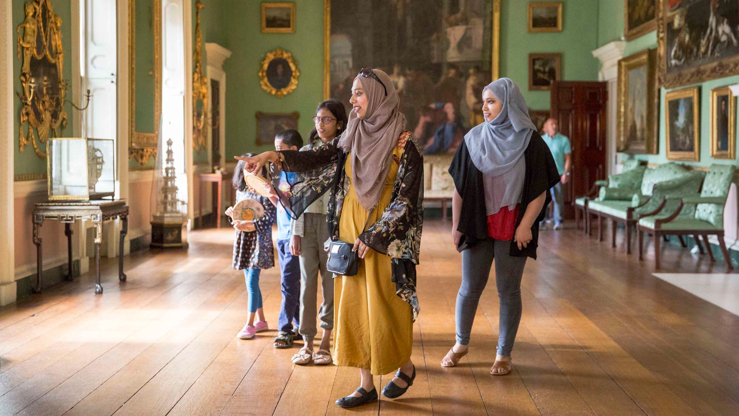 A family group are exploring the Long Gallery at Osterley House, Middlesex. The long room has walls painted green, lined with paintings in gilt frames.