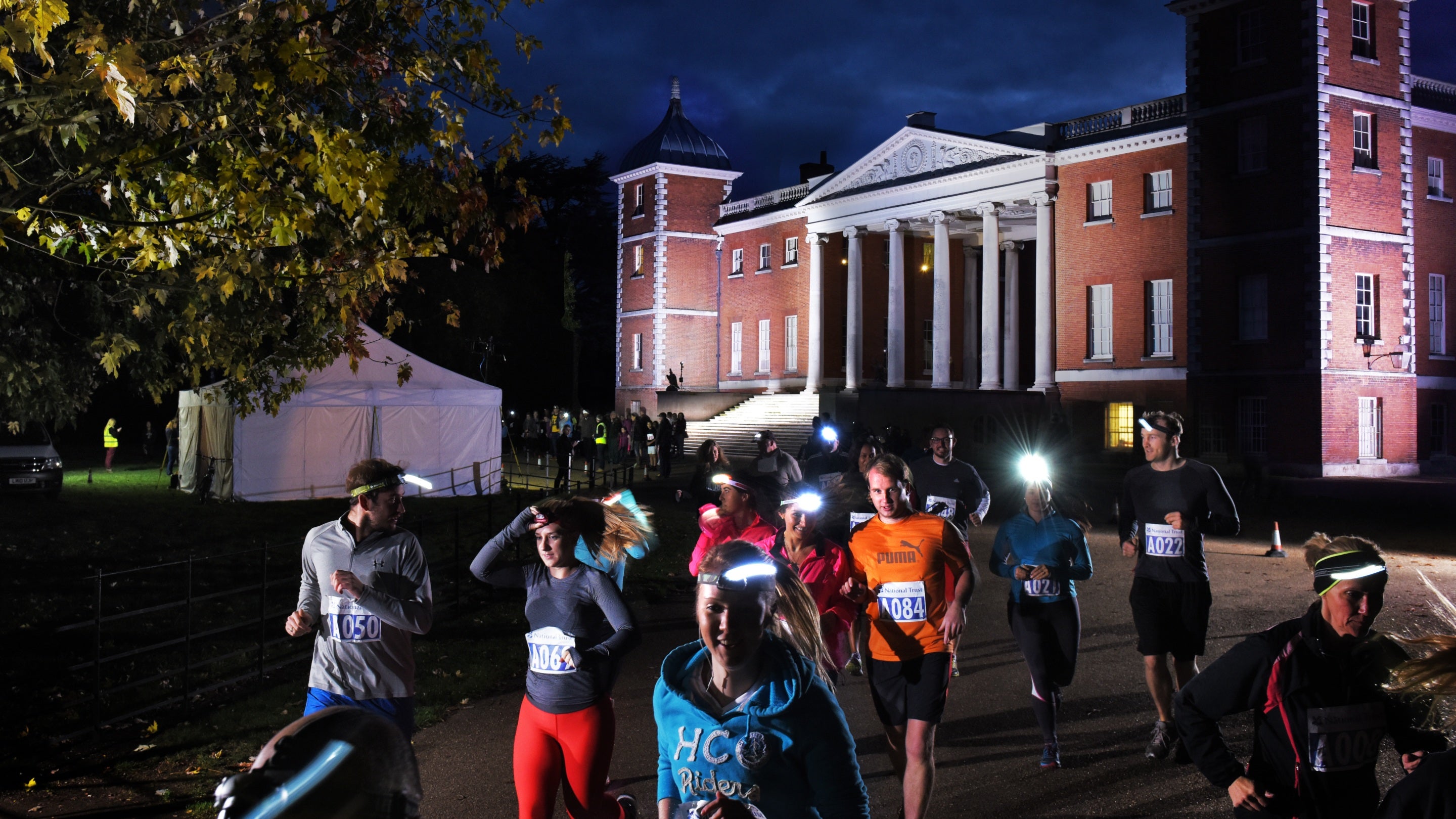 A group of runners, wearing numbers, and some wearing head torches, set off on a night run, with the house in the background, at Osterley Park and House, London