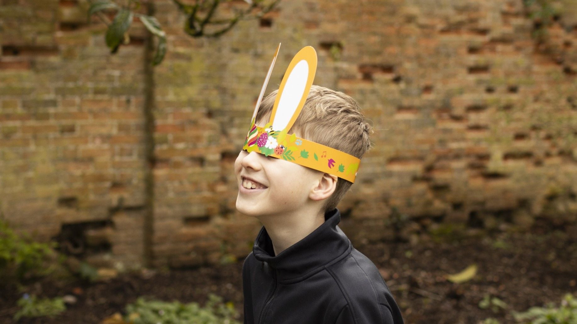 Boy in a black top wearing paper bunny ears that have slipped over his eyes with a wall in the background