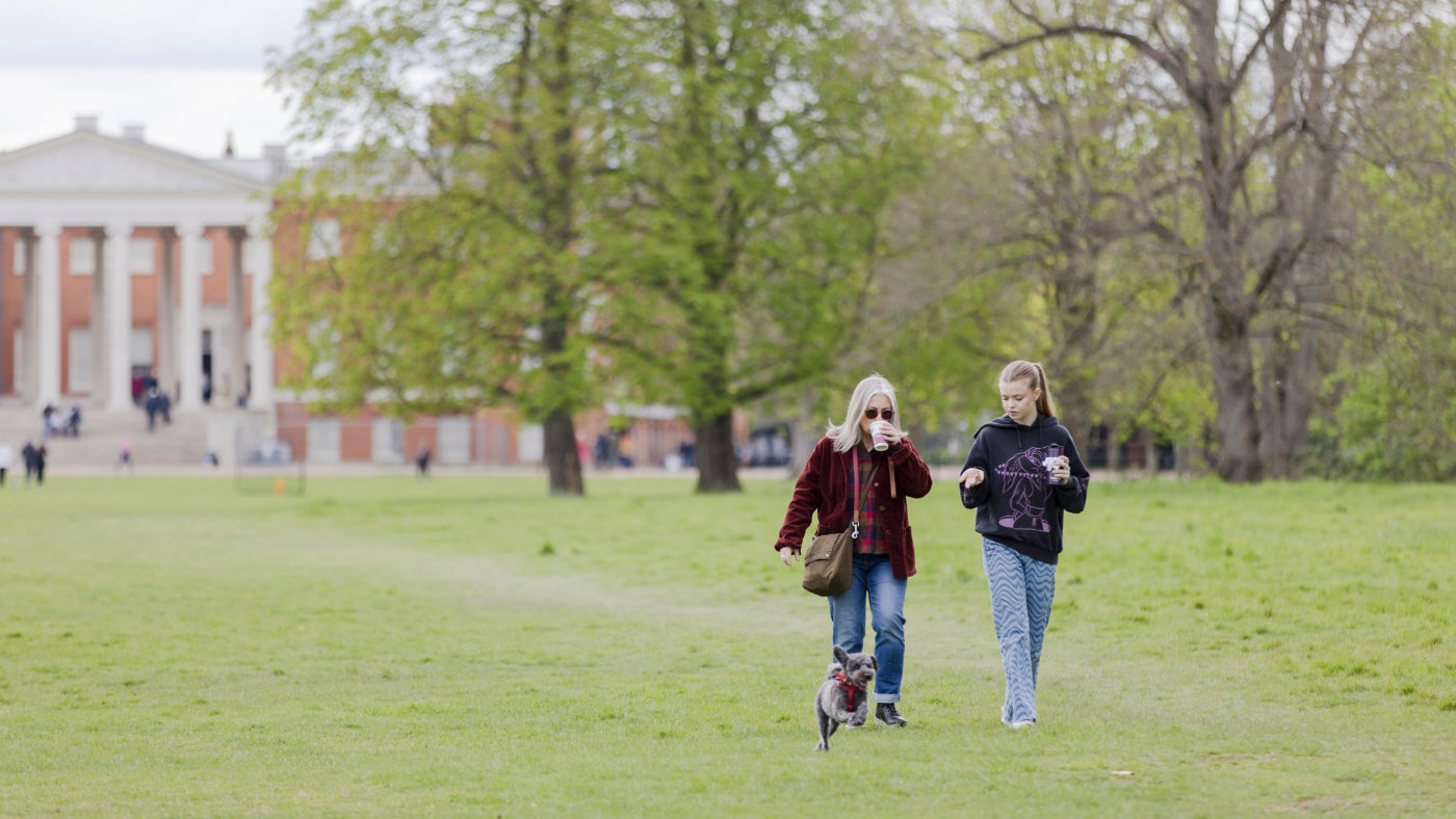 Two visitors walking away from the house at Osterley, holding cups of coffee, walking their small dog, who's jumping in the air with excitement