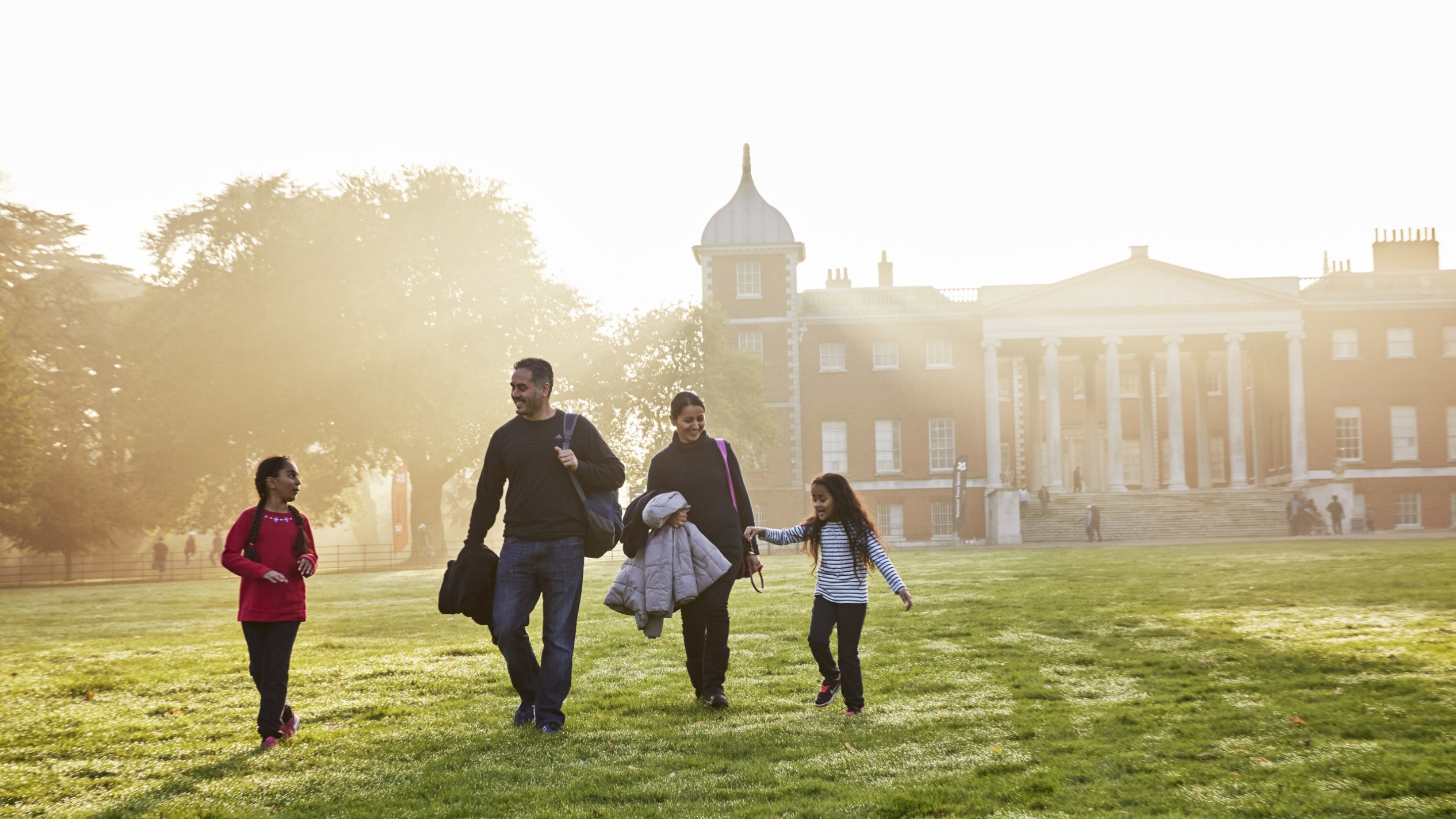 A family walking in the grounds at Osterley Park and House, London, the house is in the background and there are rays of sun coming onto the grass