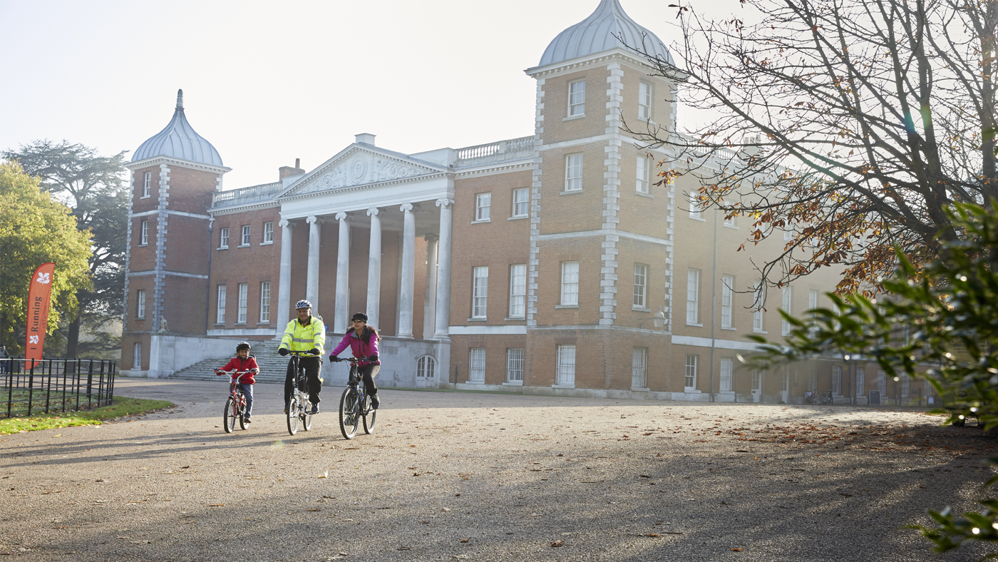 A family cycling in front of the huge façade of Osterley House, with wintry trees framing the foreground