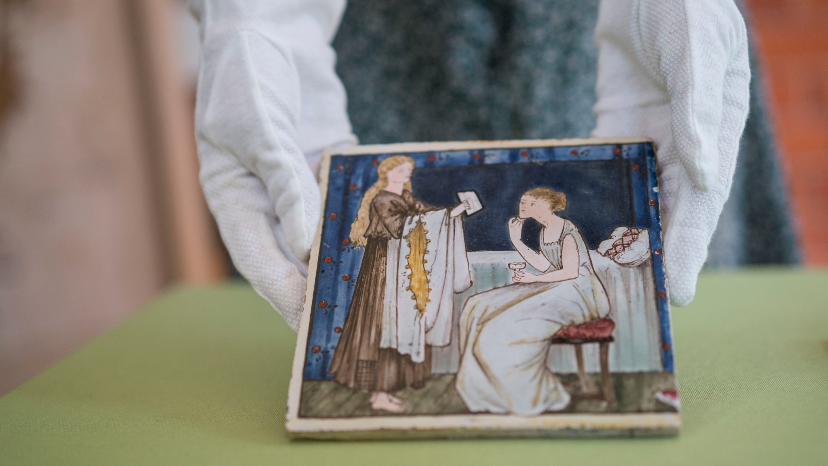 Close-up of pair of gloved hands having a tile painted with Pre-Raphaelite art