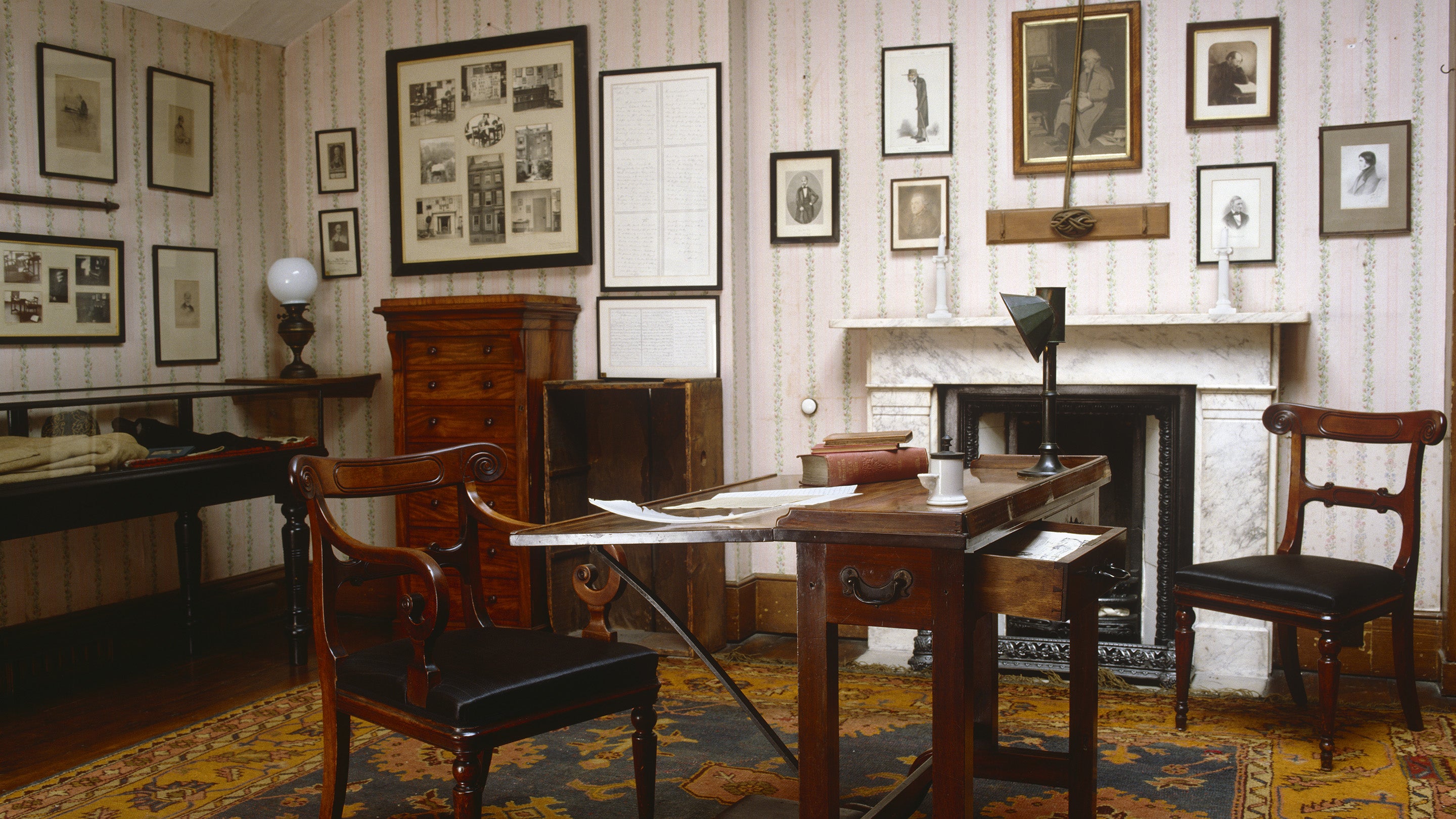 An image of the attic at Carlyle's House in London set out as it was when Carlyle wrote there using it as his study. It is a light room with a writing table and glass display case.