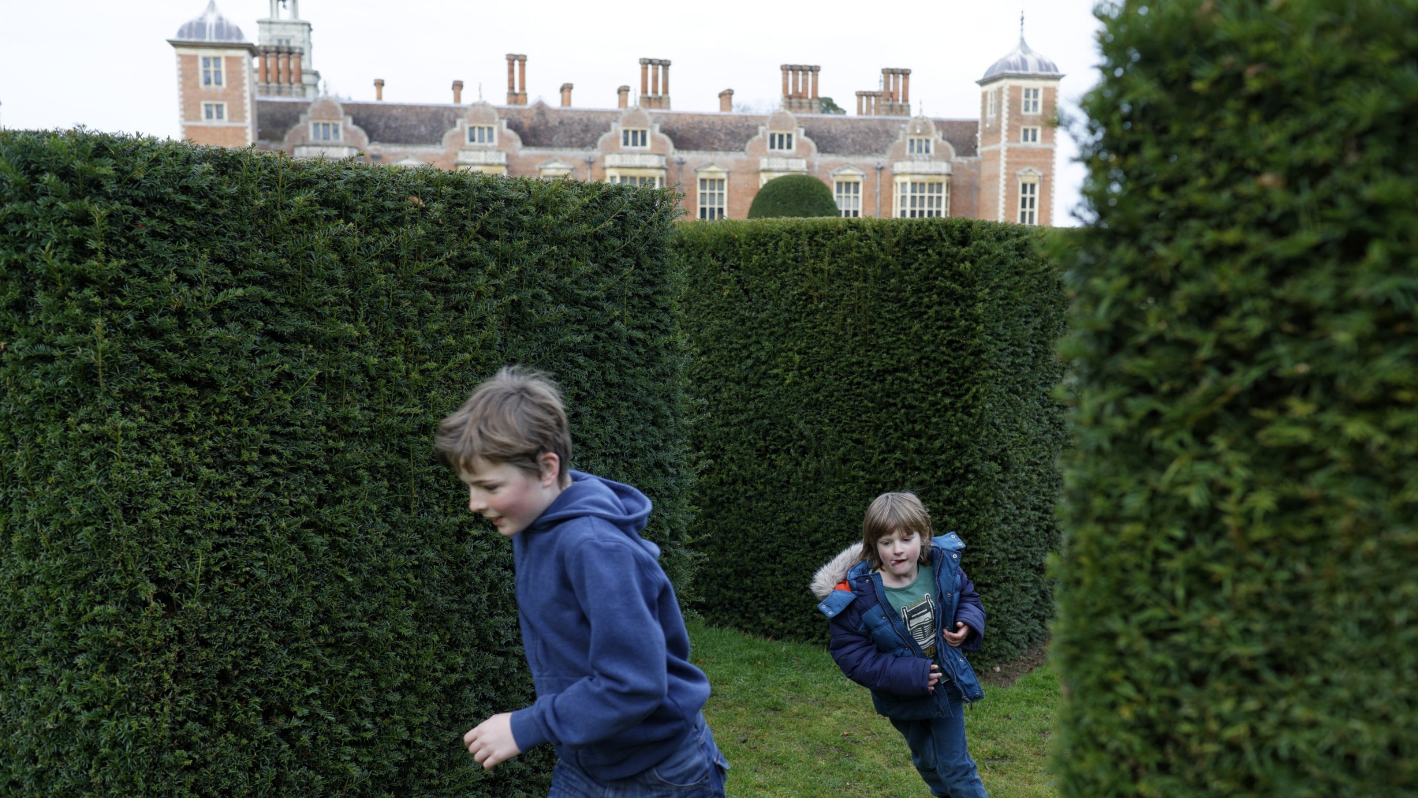 Two children dressed in blue winter wear playing amongst the clipped hedges in the grounds with Blickling Hall in the backgroound