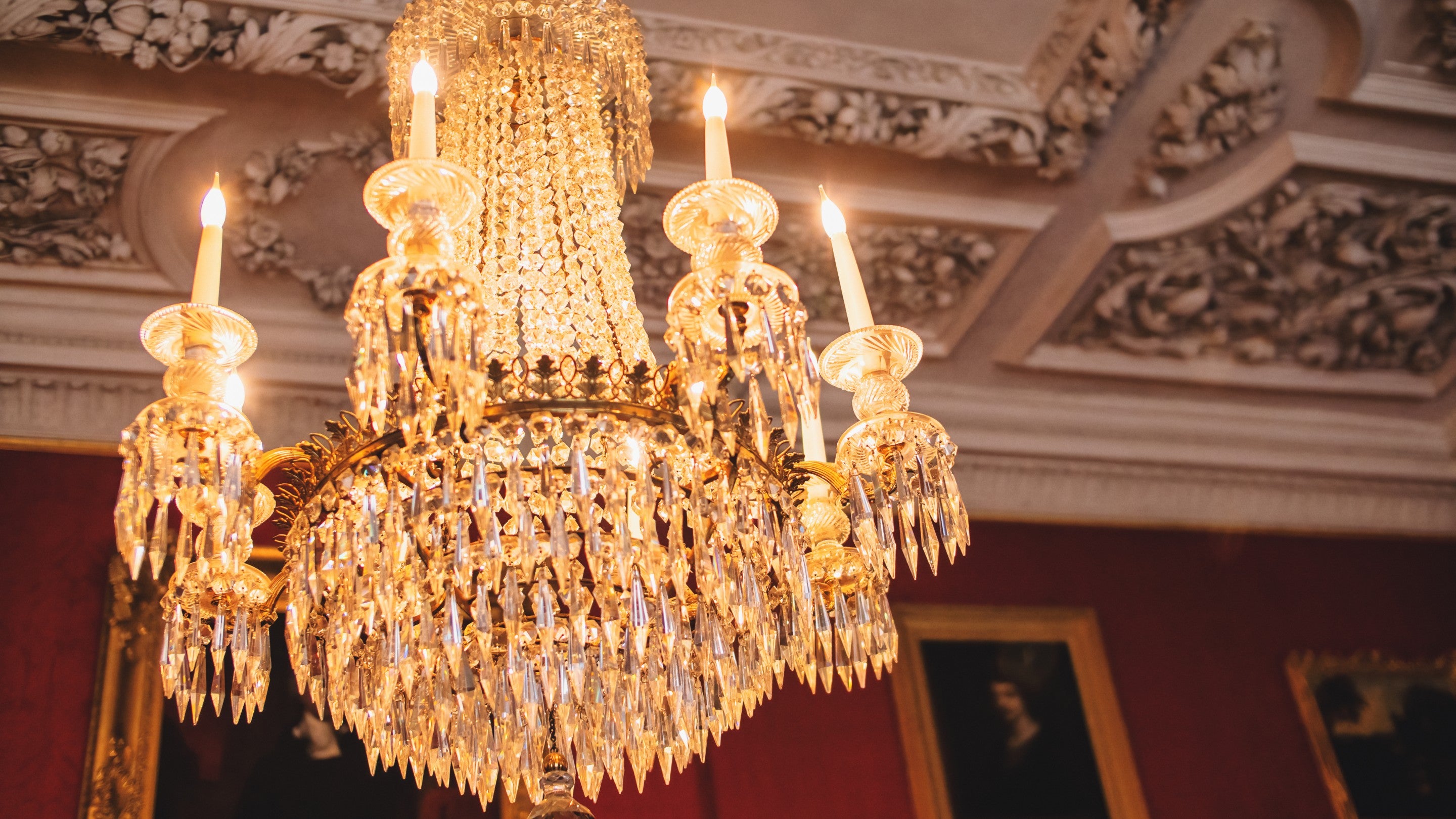 Close-up of an ornate crystal chandelier with candle-style lights hanging from a richly decorated ceiling, illuminated against a backdrop of red walls and framed portraits