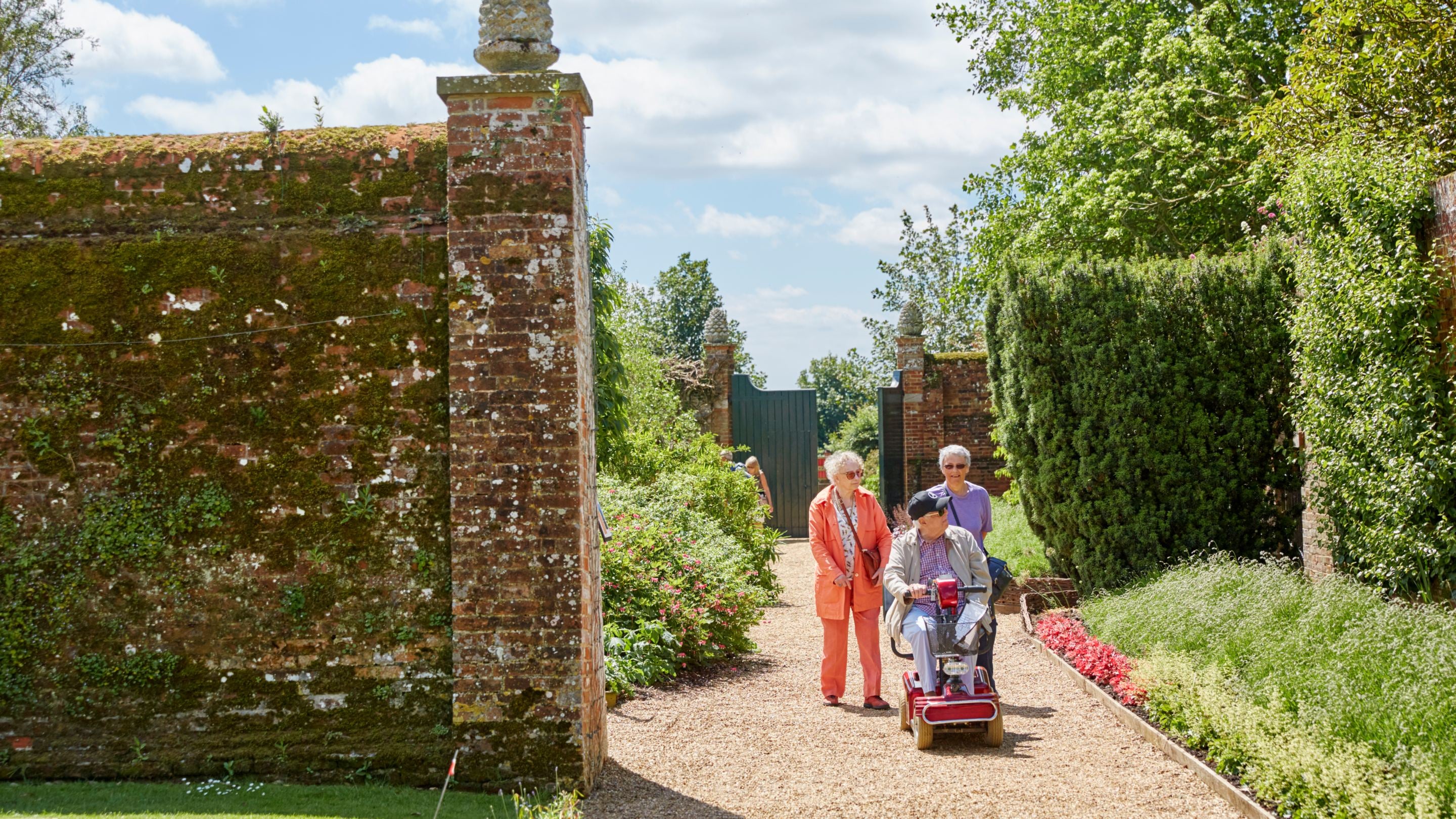 Visitors walking and using a mobility scooter on a gravel path in Felbrigg’s Walled Garden, surrounded by planting and historic brick walls.