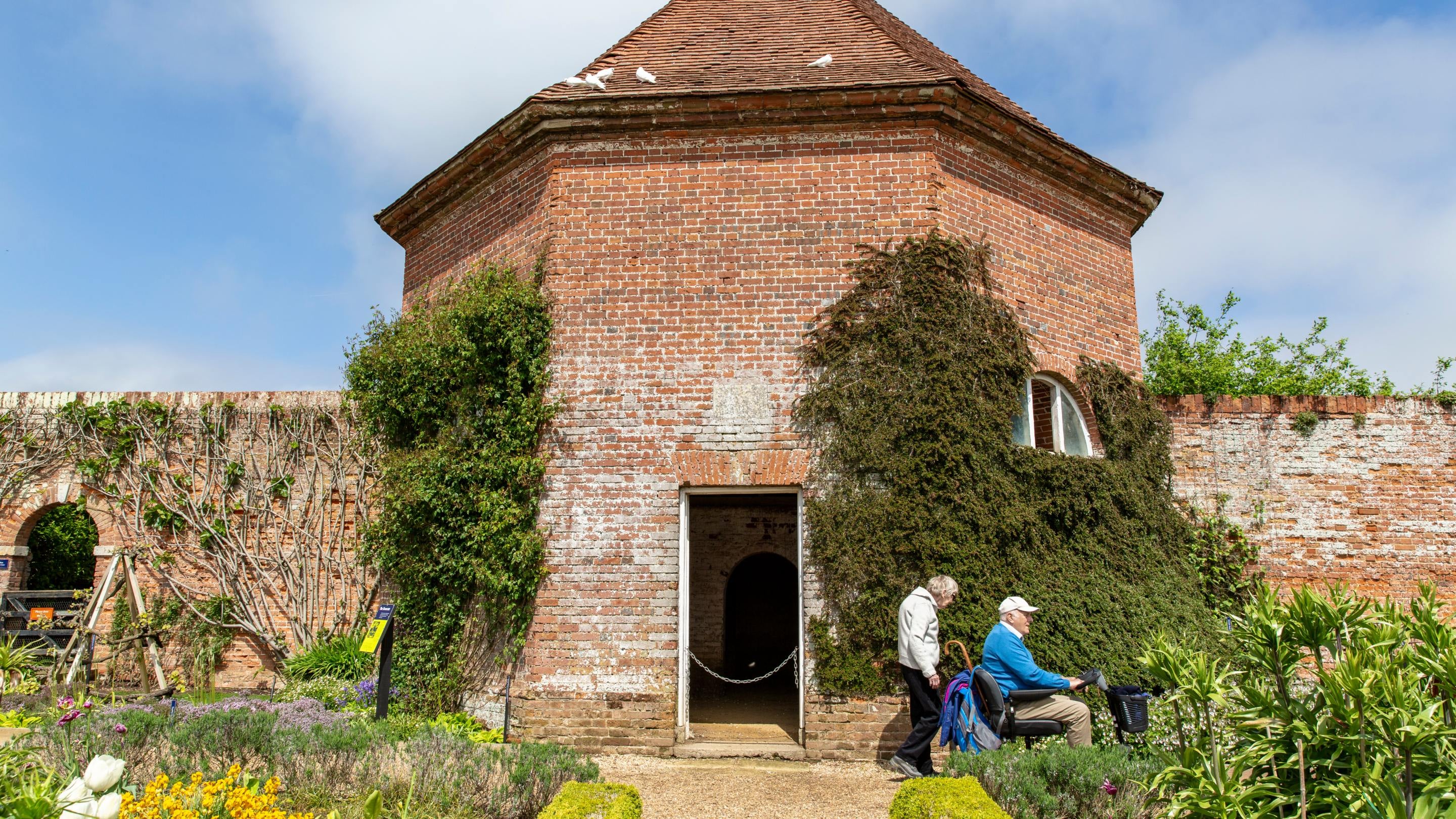 Visitors explore the Walled Garden at Felbrigg, passing a brick garden building covered in climbing plants. Colourful flower borders and neatly trimmed hedges line the path in the foreground.