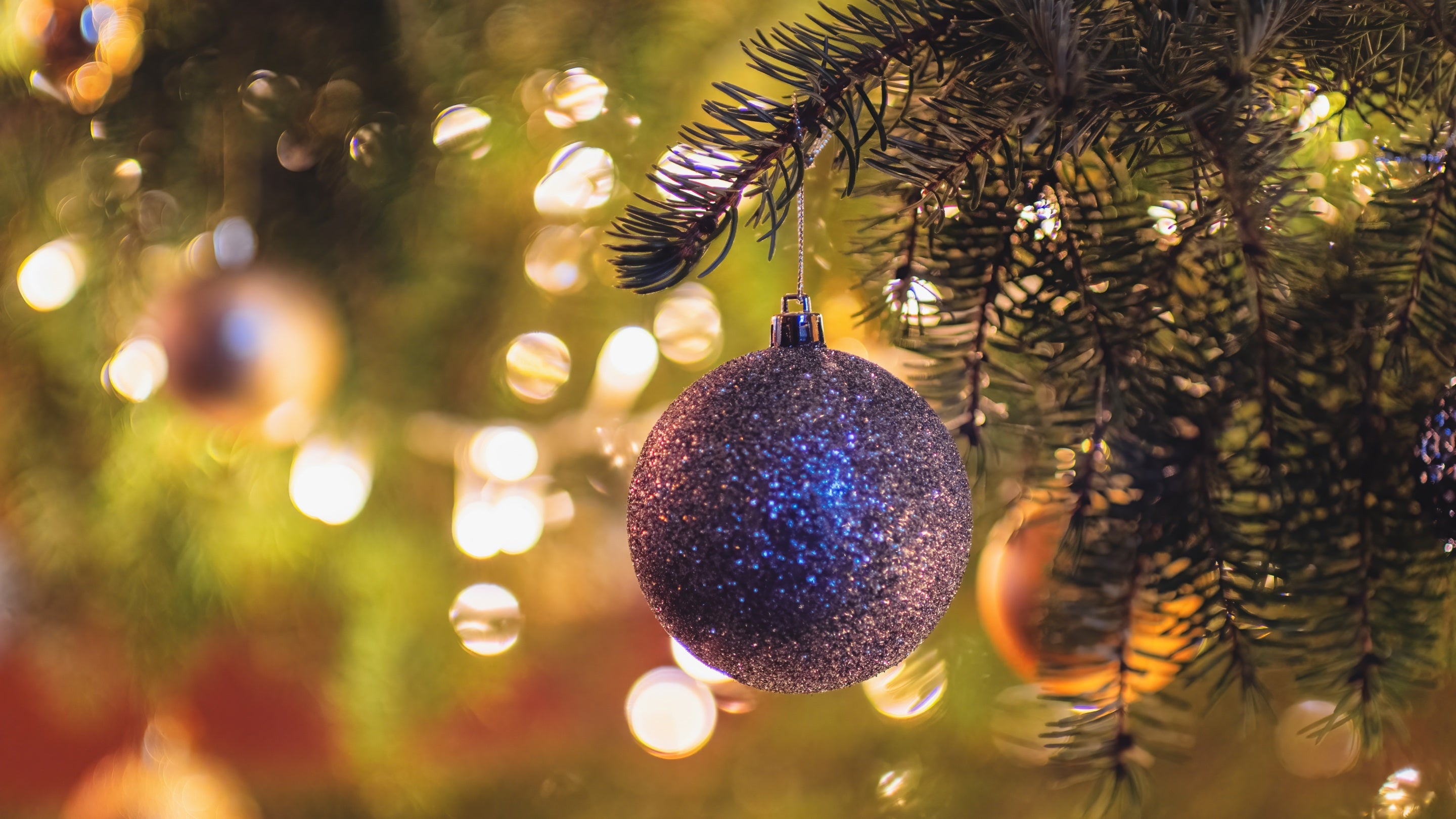 A sparkling silver bauble hangs among evergreen branches, illuminated by warm Christmas lights and festive decorations.